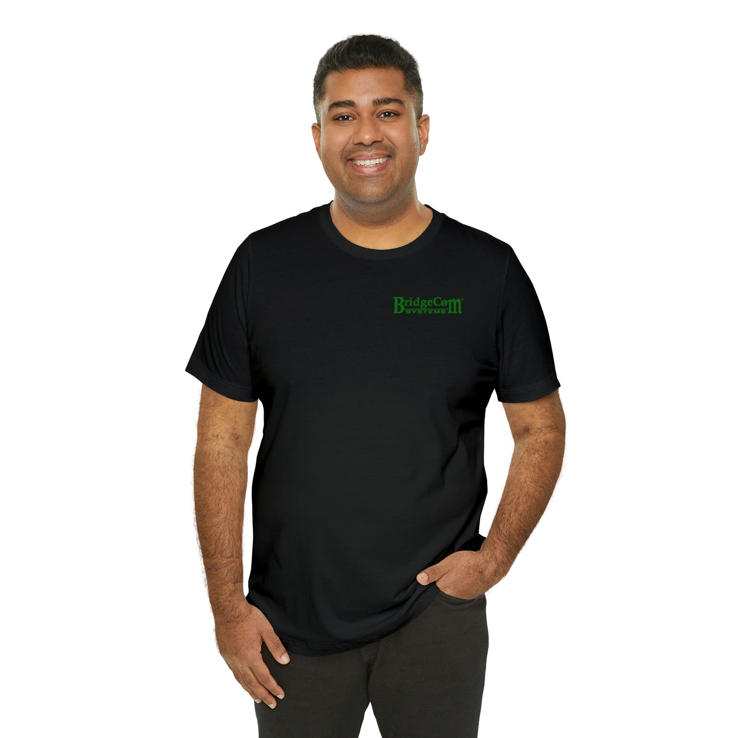 A person with short dark hair is smiling and wearing a black Short Sleeve Tee - Small Logo featuring the green BridgeCom Systems logo on the upper left side. Made from Airlume combed and ringspun cotton, their hands are resting in their pockets as they stand against a plain white background, exuding comfort and communication confidence.