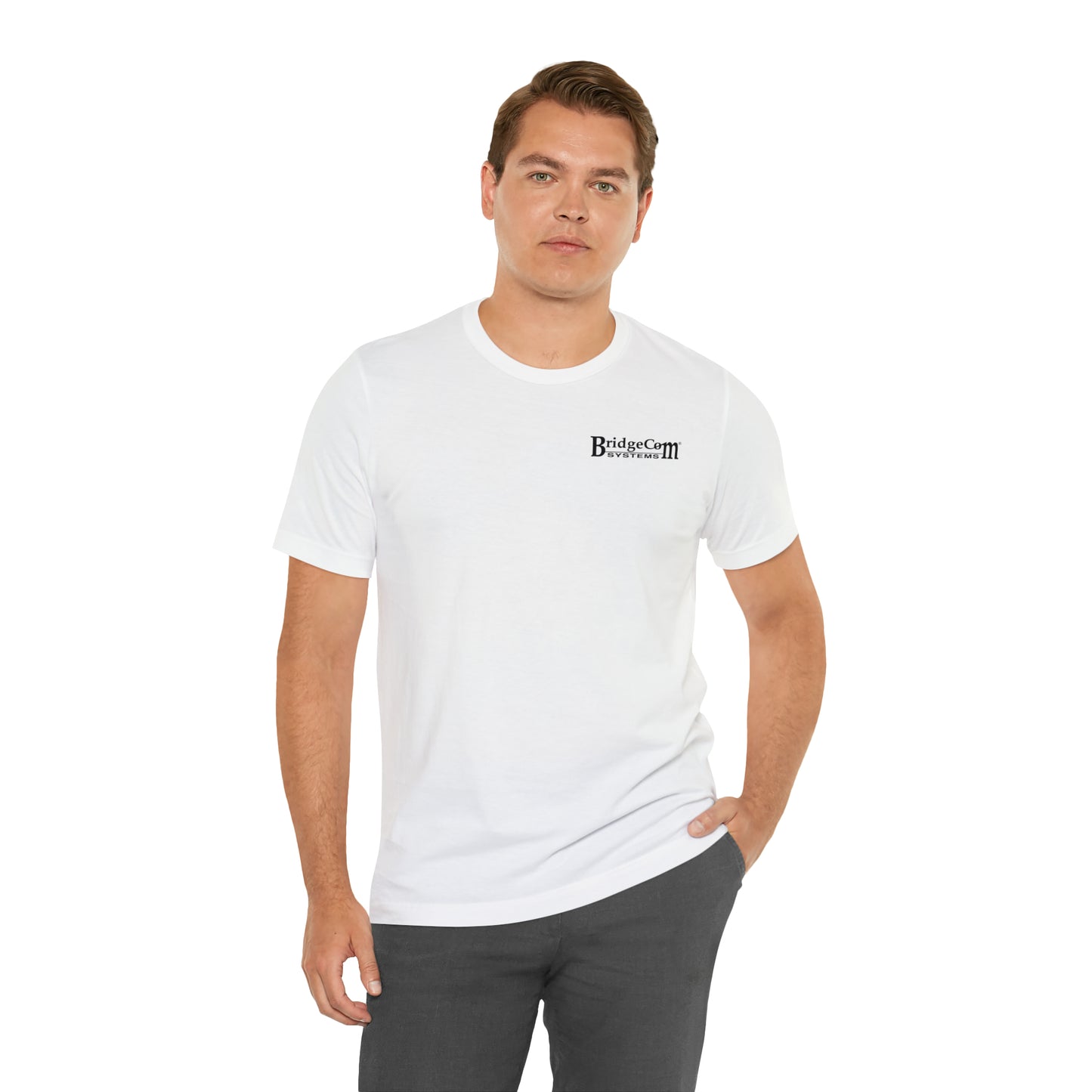 A man with short hair is wearing the Short Sleeve Tee - Small Logo, a white t-shirt featuring the BridgeCom Systems logo on the left side of his chest, paired with gray pants. He stands against a plain white background with his left hand in his pocket and maintains a neutral facial expression, embodying simplicity in communication attire.