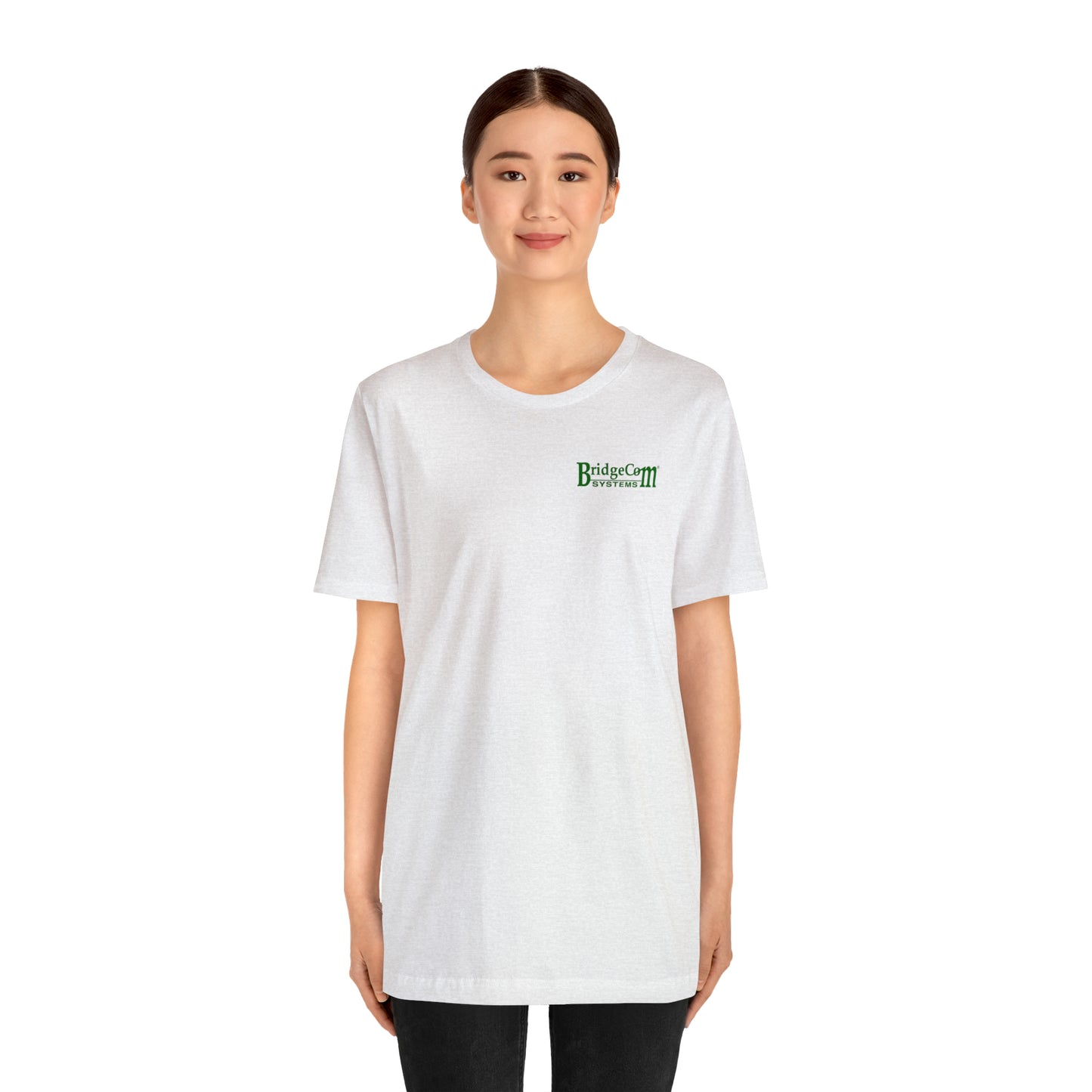 A person with dark hair tied back, wearing the Short Sleeve Tee - Small Logo from BridgeCom Systems, stands facing the camera. The white t-shirt features a small green logo that reads "BridgeCom Systems," prominently showcasing their association and emphasizing robust communication skills. The individual has a neutral expression and black pants, against a white background.