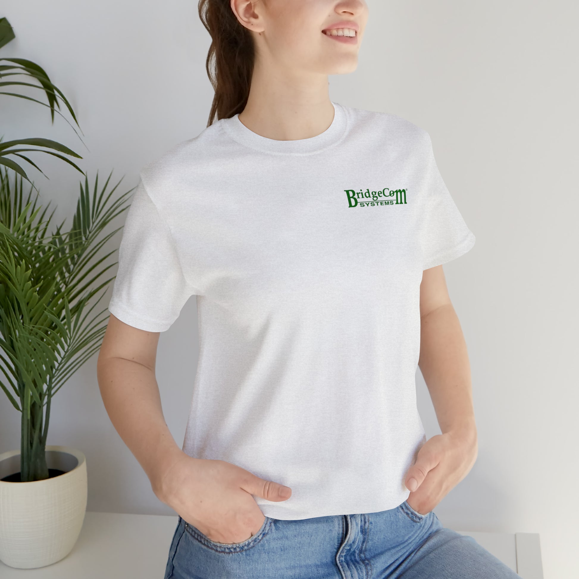 A smiling person wears the Short Sleeve Tee - Small Logo, a white t-shirt featuring the BridgeCom Systems logo in green text on the front. The person poses with one hand in their jeans pocket, embodying effortless communication. A potted plant enhances the simplicity of the background.