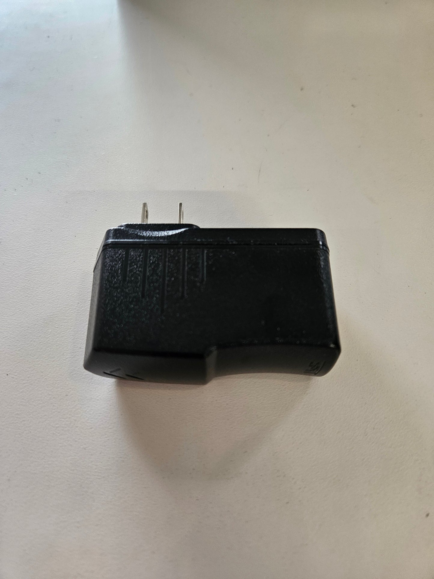 A black, rectangular 5V 3A Skybridge Power Adapter – Wall charger with upward-facing prongs lies on a flat, white surface. Its casing is adorned with textured lines and grooves, which give it a slightly worn or used appearance.