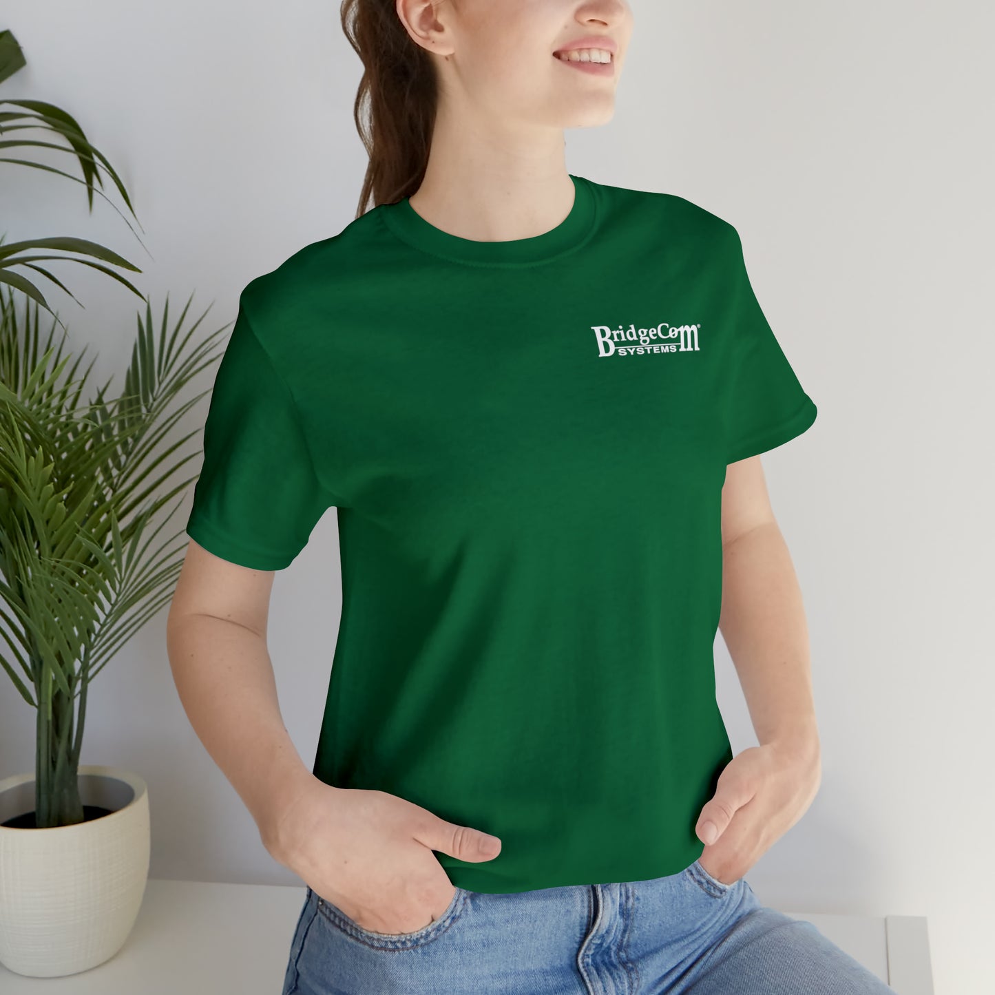 A person wearing a Short Sleeve Tee - Small Logo, made of Airlume combed cotton and featuring a green color with a "BridgeCom Systems" logo printed on the left side of the chest. They are smiling and have their hands in the pockets of their blue jeans, while a potted plant is in the background.