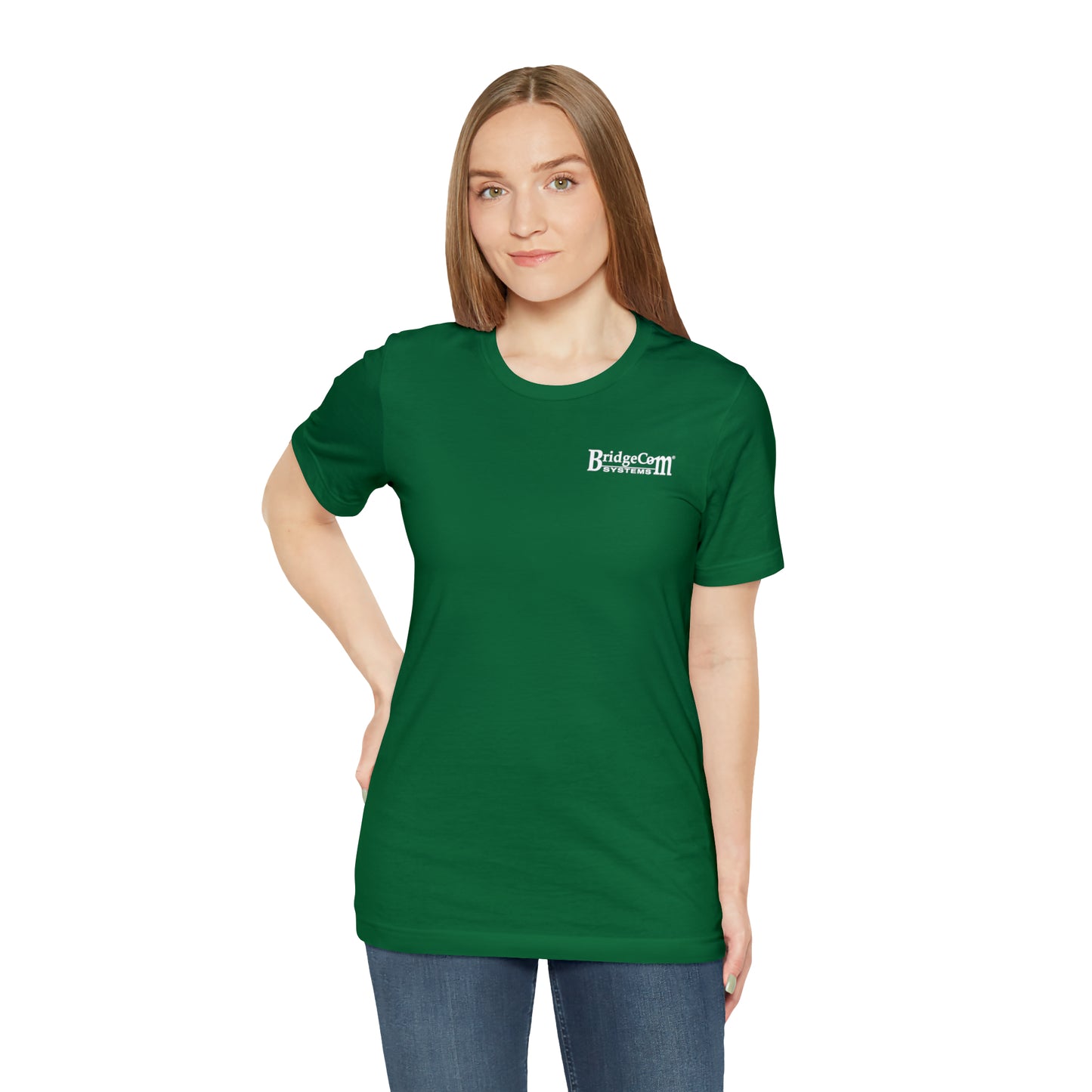 A person with long, straight hair is standing against a plain white background. They are wearing a green "Short Sleeve Tee - Small Logo" made of Airlume combed cotton and blue jeans. Their left hand is on their hip, and they are looking directly at the camera with a neutral expression.