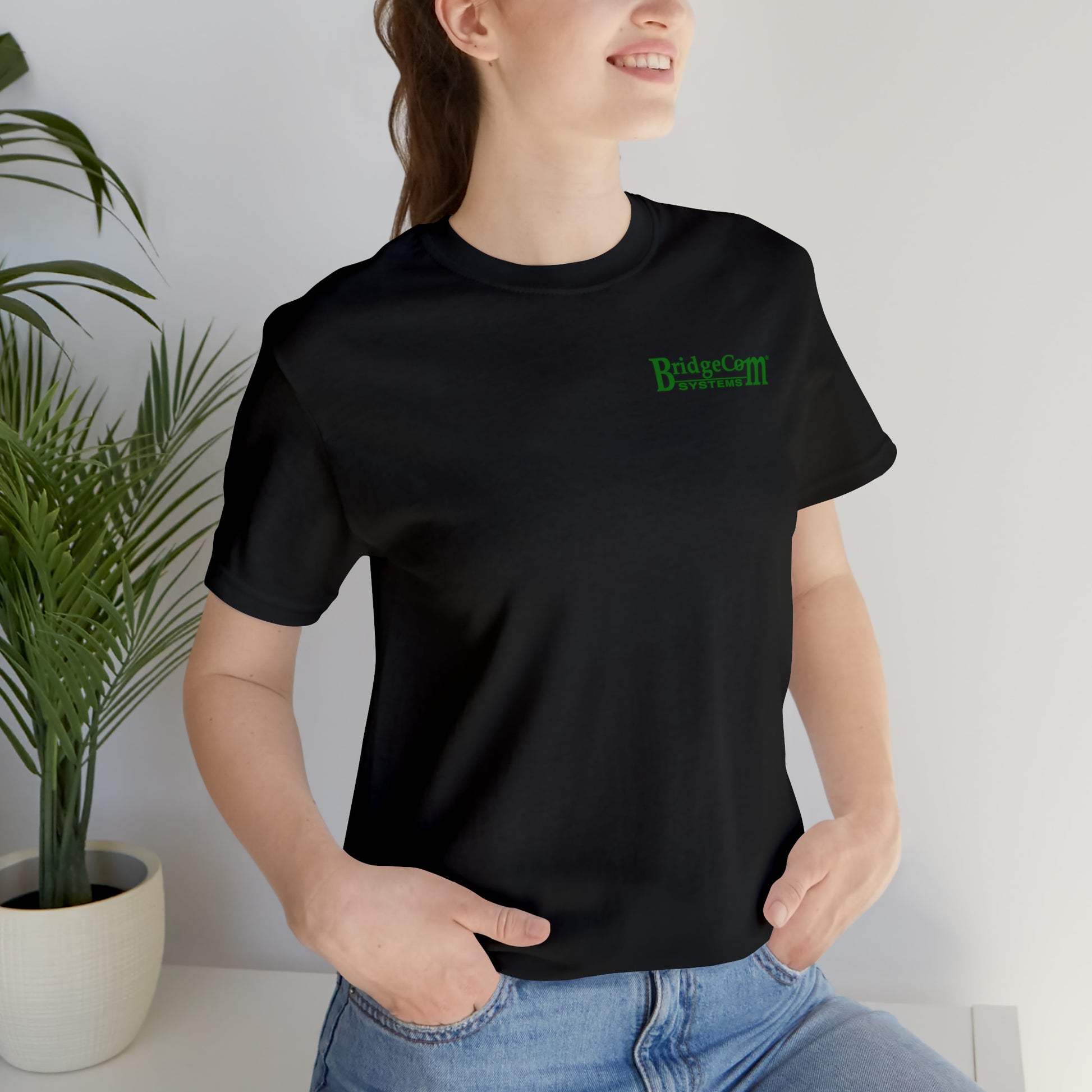A person with long hair tied back is smiling, wearing a black Short Sleeve Tee - Small Logo from BridgeCom Systems with green text. With one hand in their jean pocket, they stand next to a potted plant against a light-colored wall, embodying the spirit of effective communication.