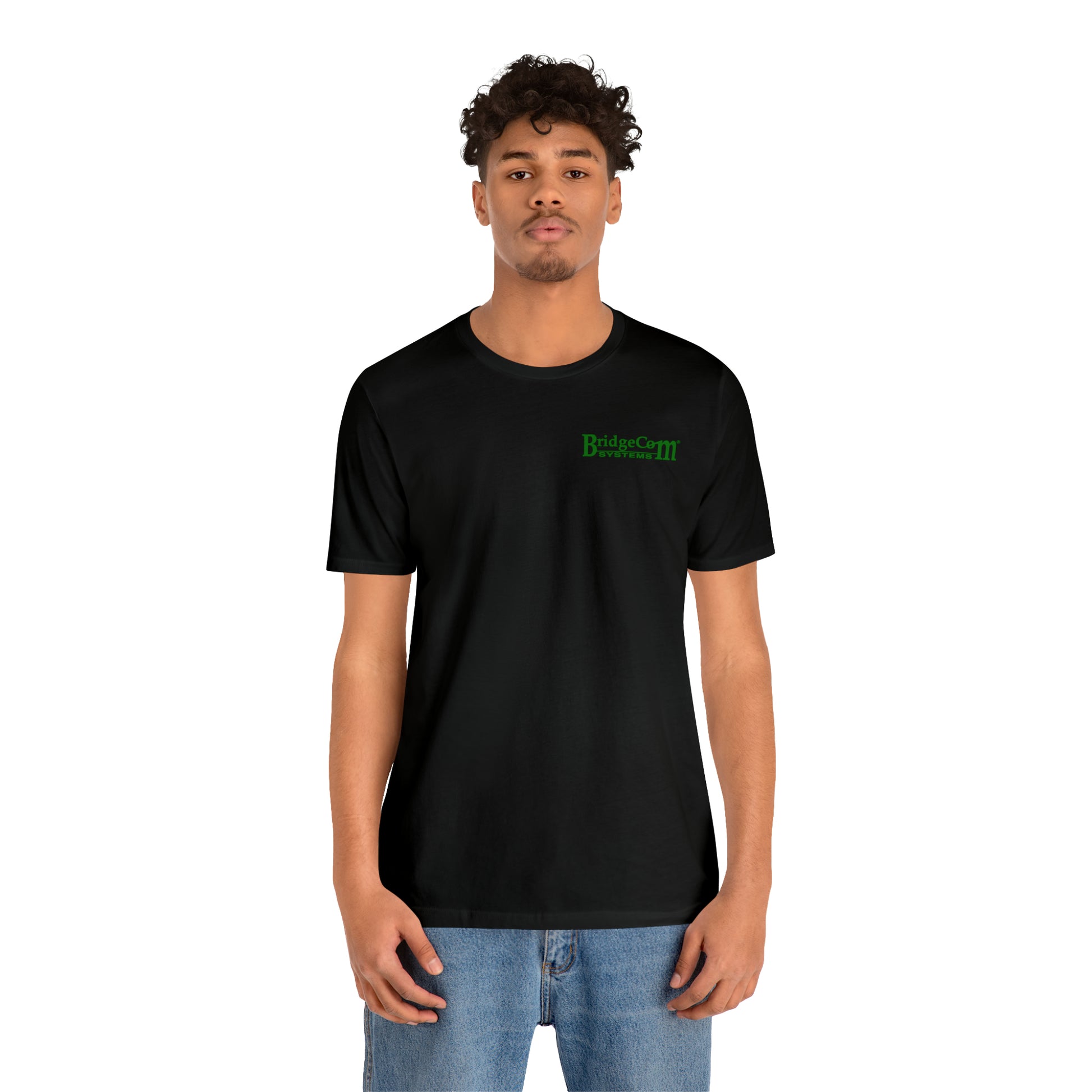 A young man with curly dark hair and light blue jeans stands against a white background, wearing a Short Sleeve Tee - Small Logo, made of soft Airlume cotton. The black T-shirt features green text that reads "Bridgecom Systems.