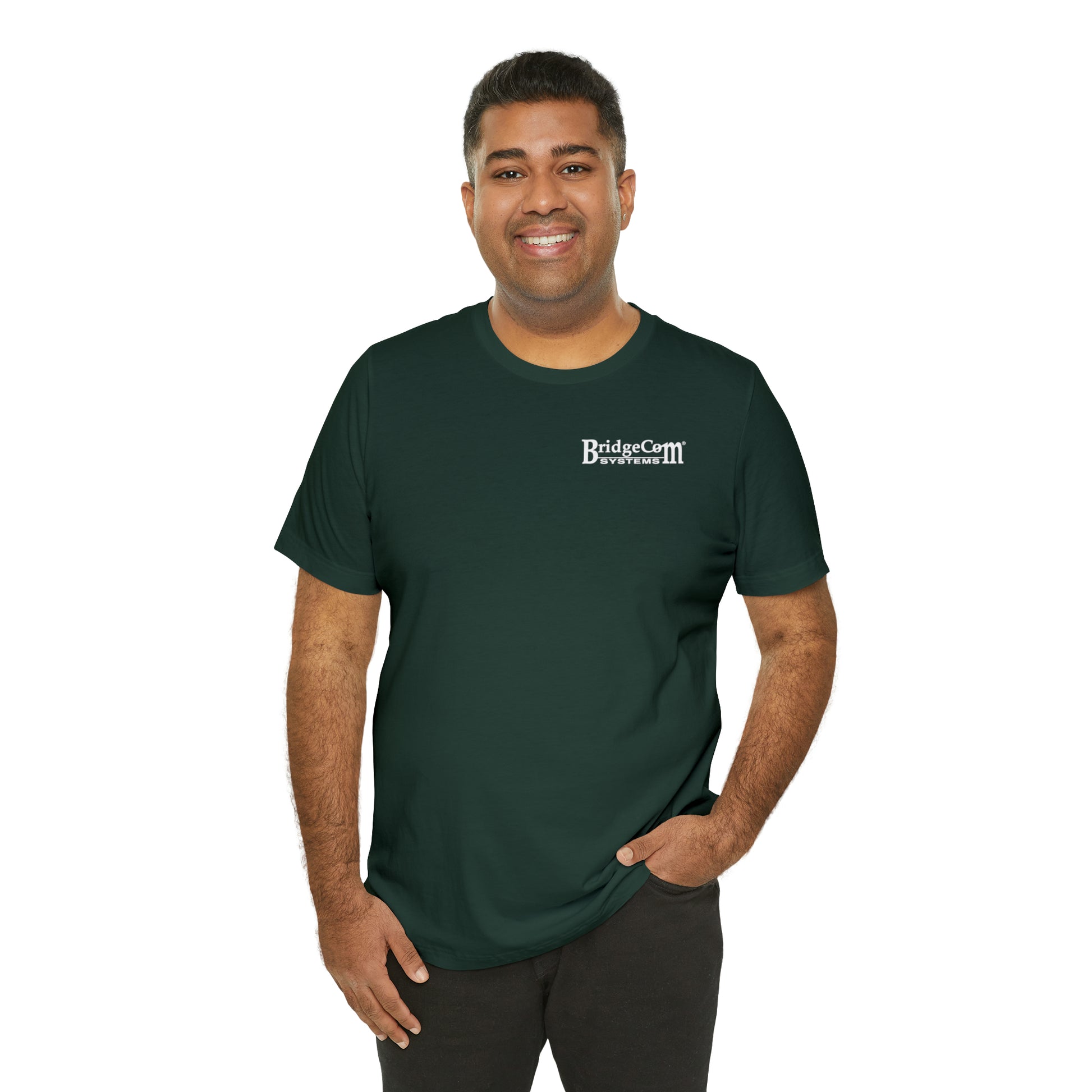 A smiling man is standing and wearing a dark green "BridgeCom Systems" short sleeve tee made from soft Airlume cotton. He has short dark hair and is posing with one hand in his pocket against a plain white background, proudly displaying the small BridgeCom Systems logo.