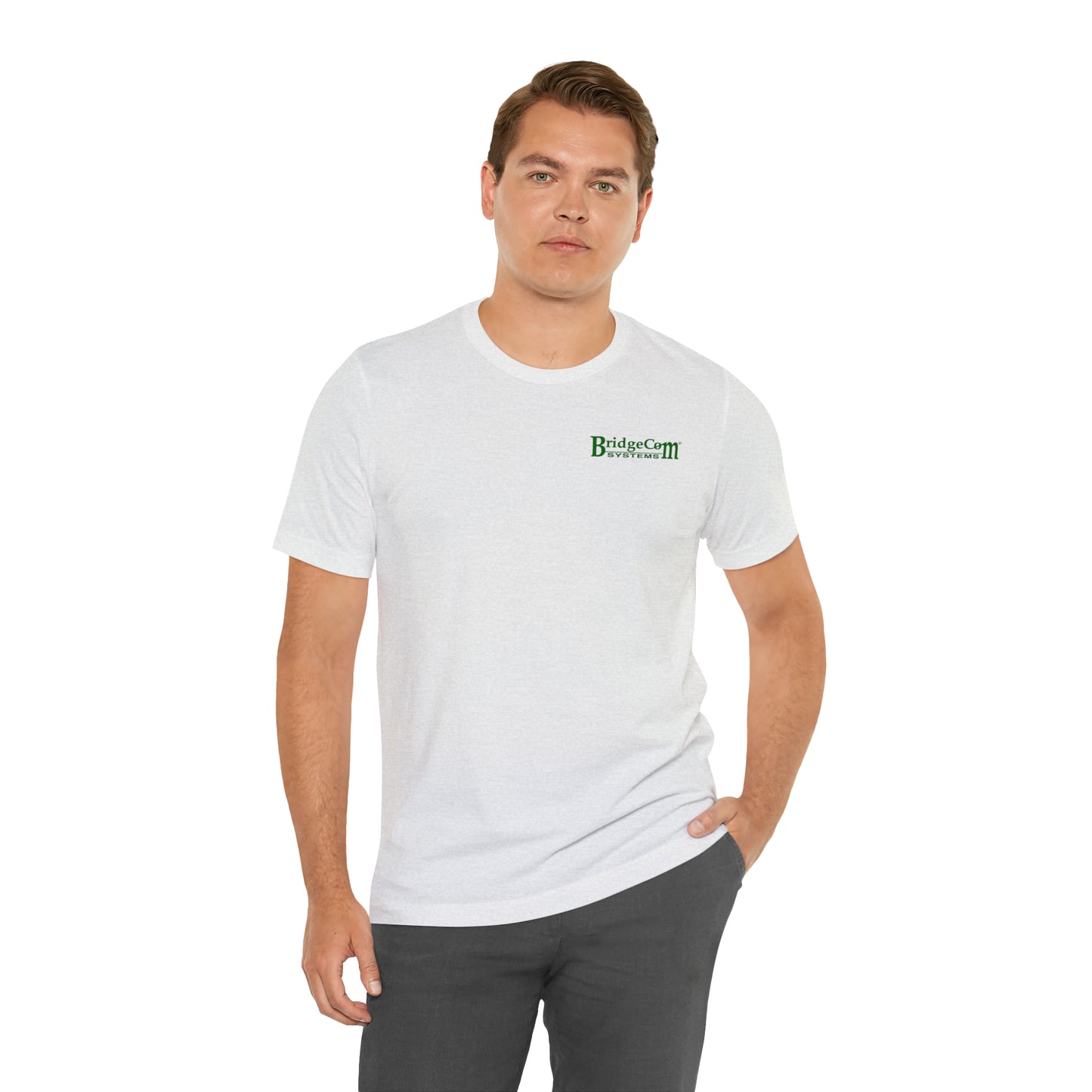 A man with short hair stands against a plain white background, wearing a "Short Sleeve Tee - Small Logo" made of 100% Airlume cotton, which features a small green and black "BridgeCom Systems" logo on the left chest. He pairs it with gray pants, has his left hand in his pocket, and maintains a neutral expression.