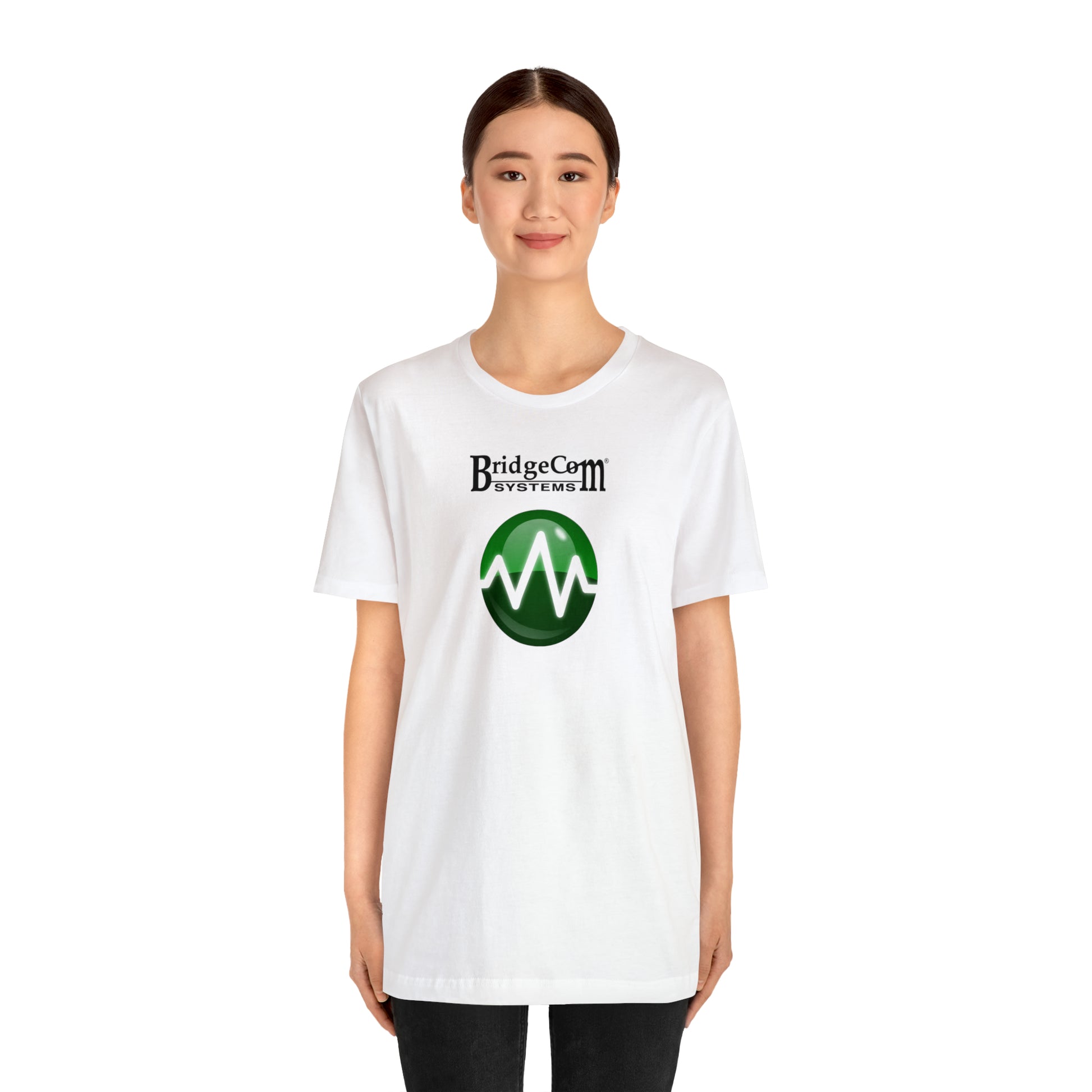 A person with long dark hair tied back is standing against a plain background, wearing a Unisex Short Sleeve BridgeCom Tee - Alternate Logo featuring the text "BridgeCom Systems" above a green circular logo with a white zigzag line. They are facing forward and smiling slightly.