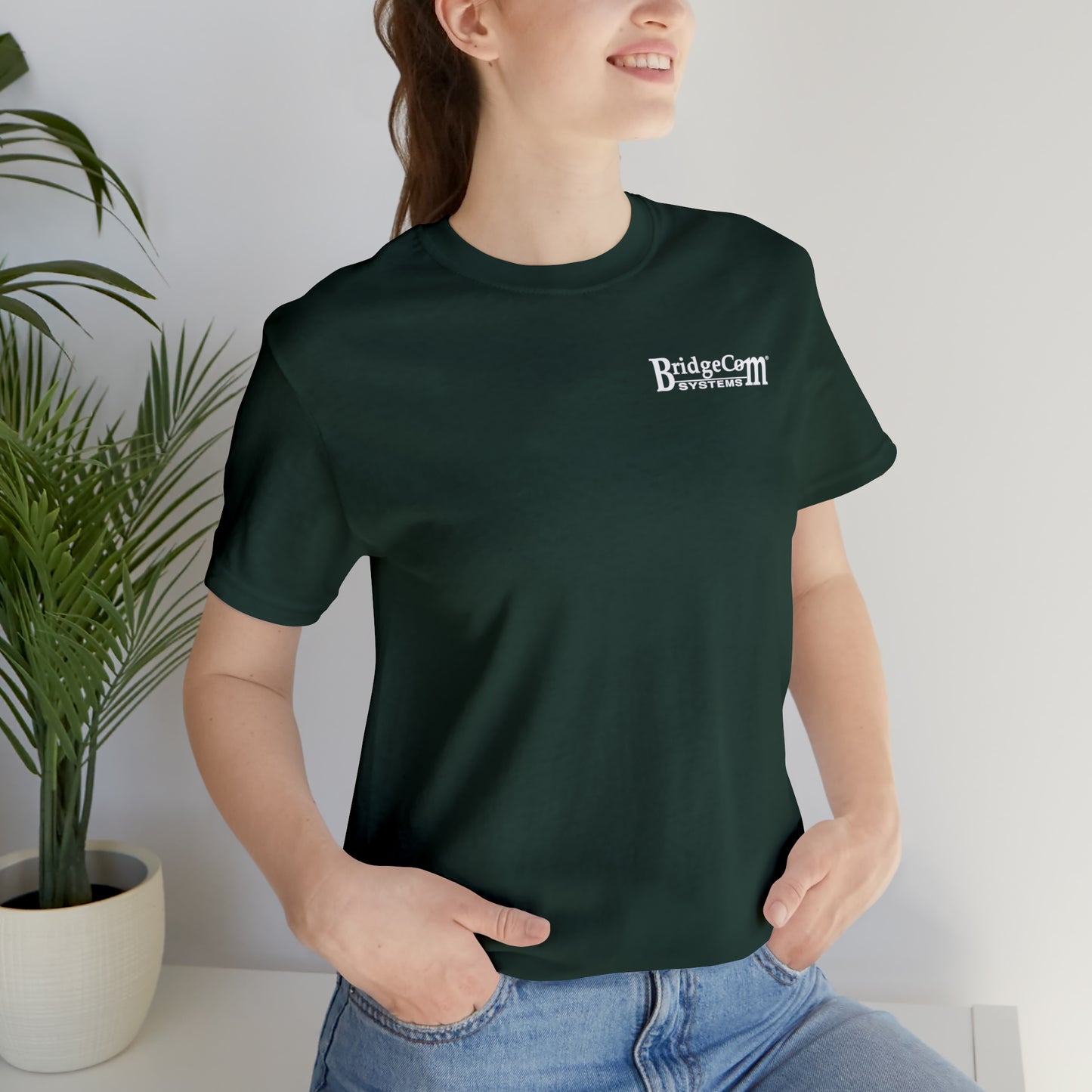 A person with long brown hair, tied in a ponytail, is wearing a dark green Short Sleeve Tee - Small Logo made of Airlume cotton with the BridgeCom Systems logo printed in white on the left chest area. They are smiling, standing next to a potted plant, and have both hands resting in the pockets of their blue jeans.