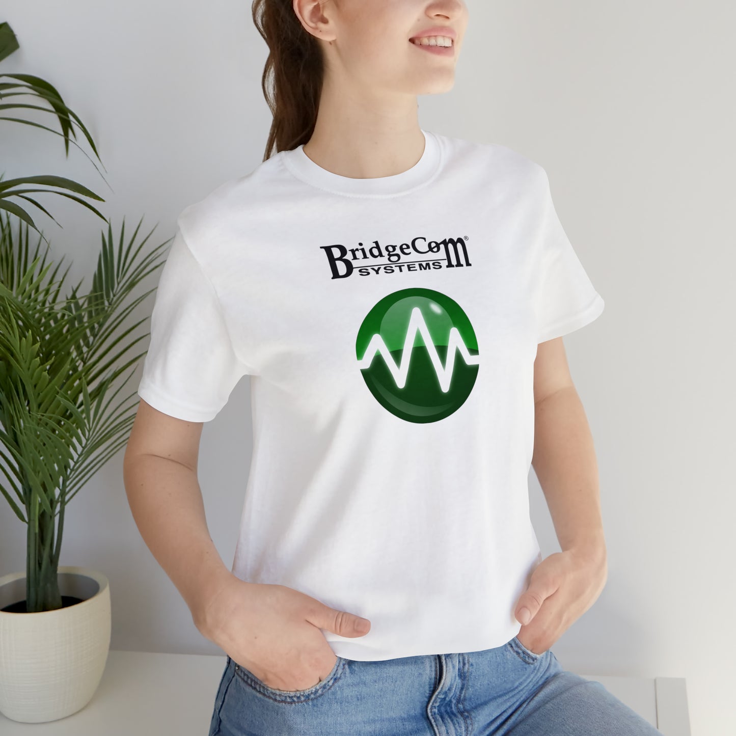 A person wearing a white Unisex Short Sleeve BridgeCom Tee - Alternate Logo made from soft Airlume cotton, featuring a green circular design with a white waveform, stands near green potted plants. Smiling with their hands in their jean pockets, they embody the essence of modern communication.