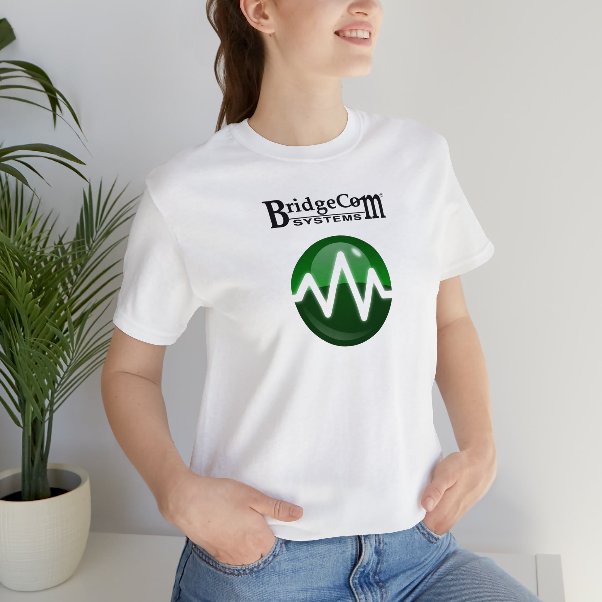 A person wearing a white Unisex Short Sleeve BridgeCom Tee - Alternate Logo made from soft Airlume cotton, featuring a green circular design with a white waveform, stands near green potted plants. Smiling with their hands in their jean pockets, they embody the essence of modern communication.