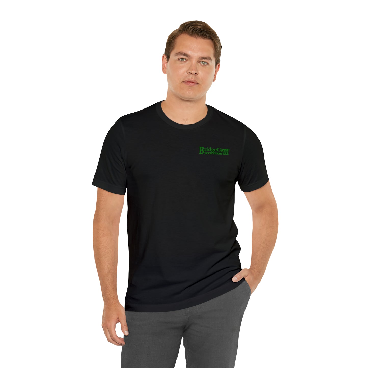 A person with short, light brown hair stands with one hand in their pocket and the other by their side. They are wearing a black "Short Sleeve Tee - Small Logo" made of Airlume cotton, featuring green text that reads "BridgeCom" on the left side of the chest, along with gray pants. The background is white.
