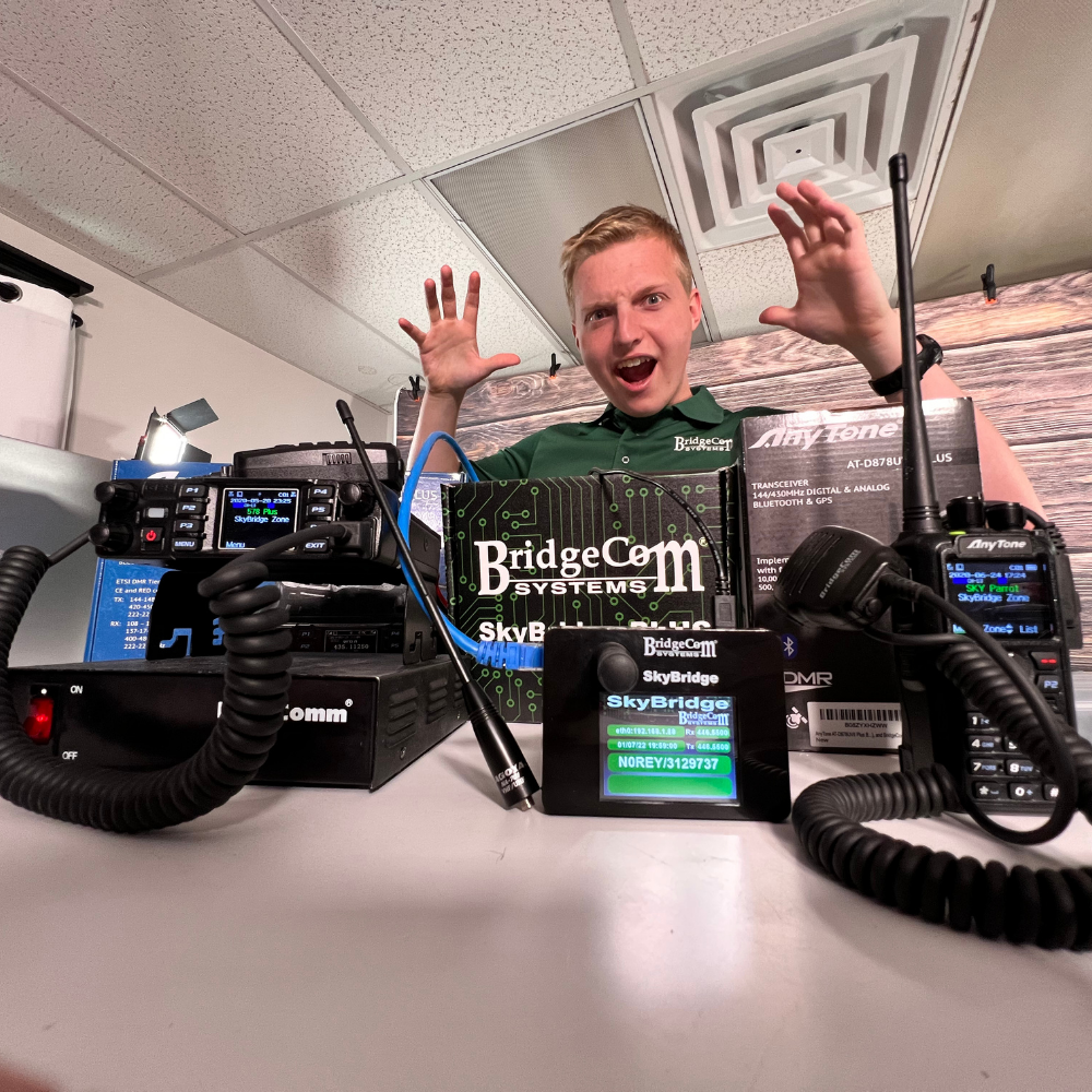 A smiling person raises their hands behind a table displaying the Ultimate Hamshack Pack with $291 in Training Course FREE!, featuring advanced DMR technology and products from BridgeCom Systems and SkyBridge under bright indoor lighting.