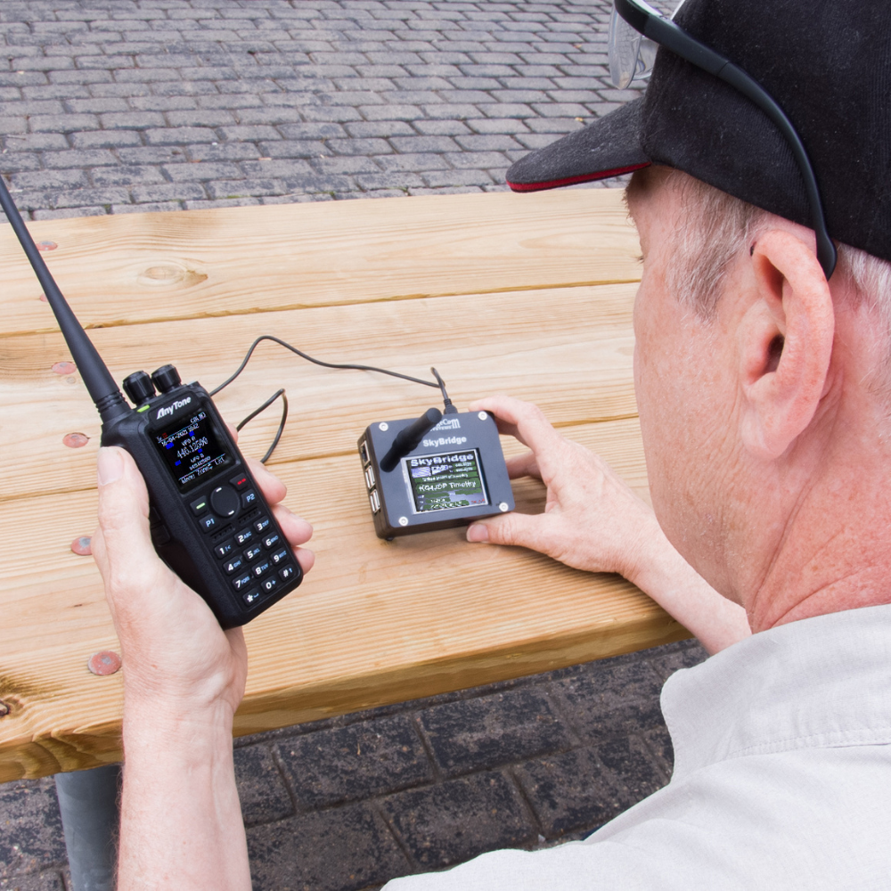 Outdoors on a wooden bench, someone holds a ham radio in one hand and the SkyBridge MAX Plug and Play Upgrade with display in the other; both devices are connected by a cable.