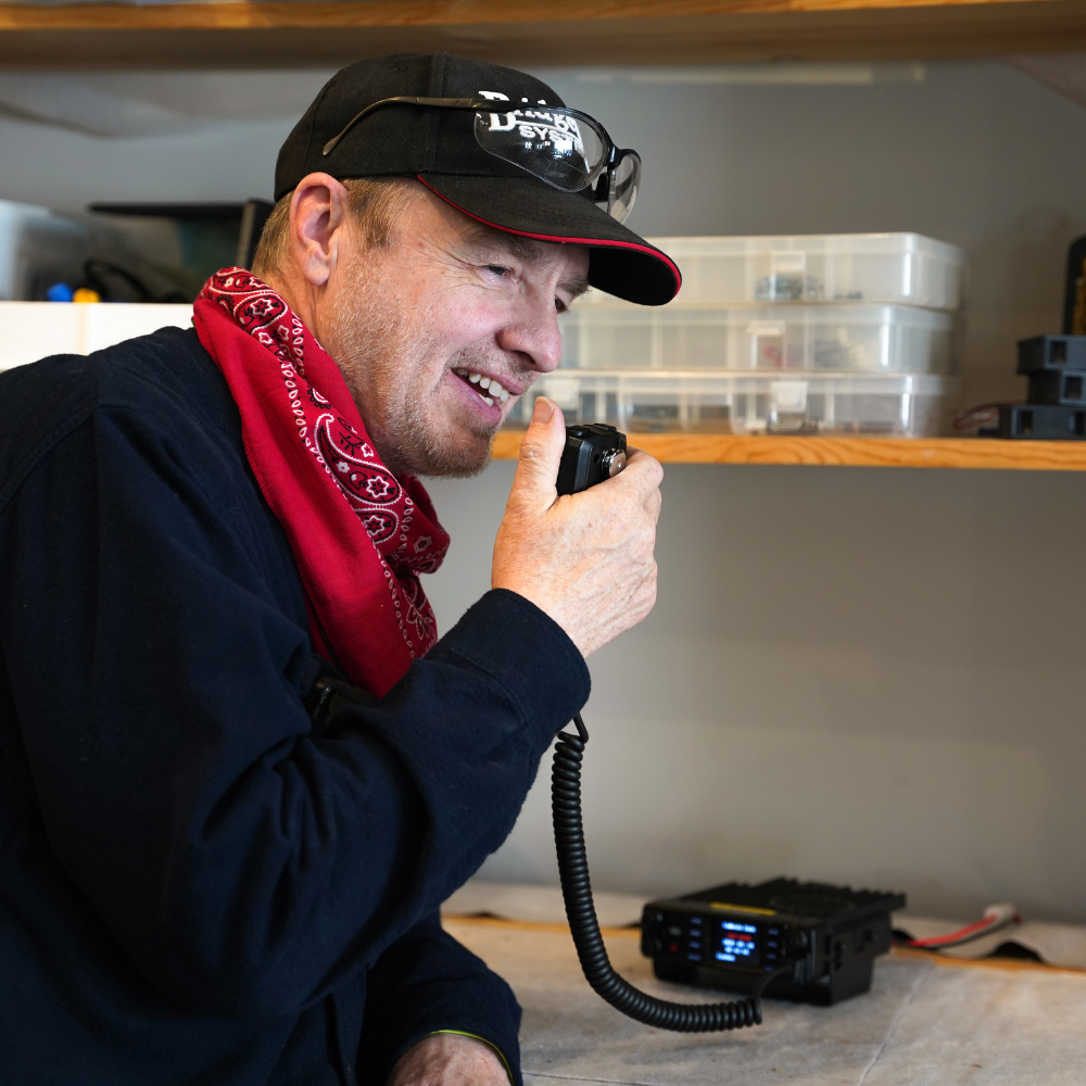 A man in a black cap, red bandana, and dark shirt speaks into a handheld radio in a workshop, with plastic storage boxes and a Mobile Plug and Play Package: AnyTone AT-D578UVIII PLUS Mobile Radio w/ SkyBridge Max Hotspot on the shelf behind him.