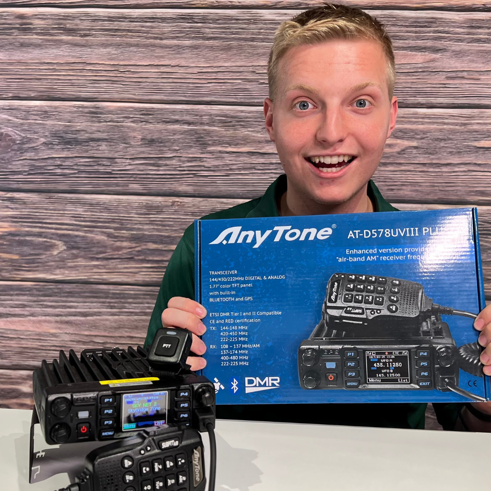 With excitement, a person holds the blue AnyTone 578UVIII PLUS box while admiring the Tri-Band Mobile Radio supporting DMR and analog on the table. The background's wood texture adds warmth to the scene.