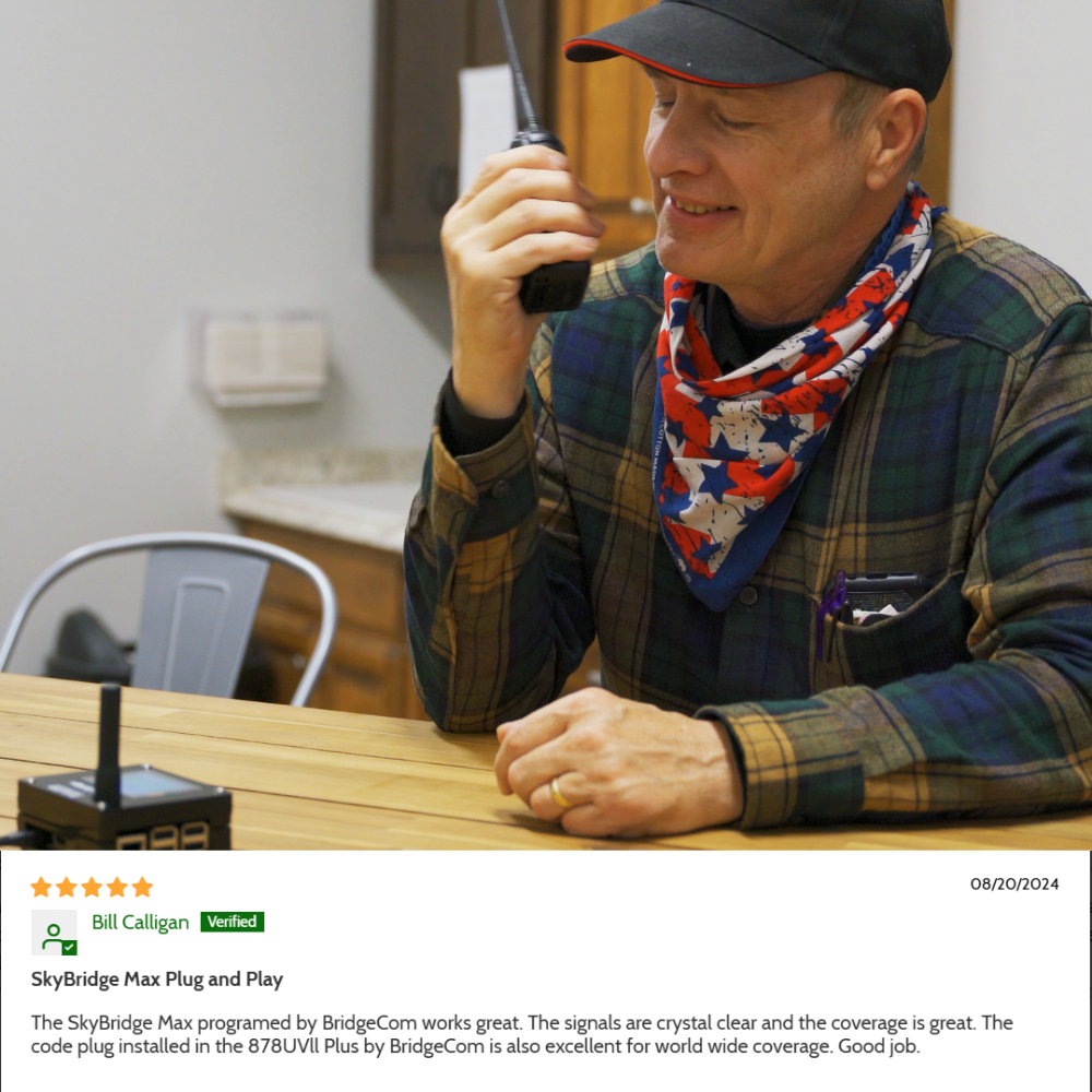 A smiling man in a red and blue scarf and cap sits at a table, talking into a handheld ham radio. On the table are electronic devices, including the SkyBridge MAX Plug and Play Upgrade hotspot.