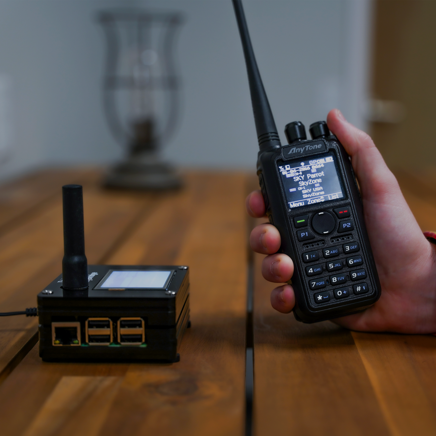 A person holds an AnyTone AT-D878UVII Plus with its screen showing menu options. Nearby, a small black device with an antenna and ports sits on a wooden table. A blurred decorative item enhances the depth of this dual band DMR/Analog handheld setup, complete with a $97 training course for free!.