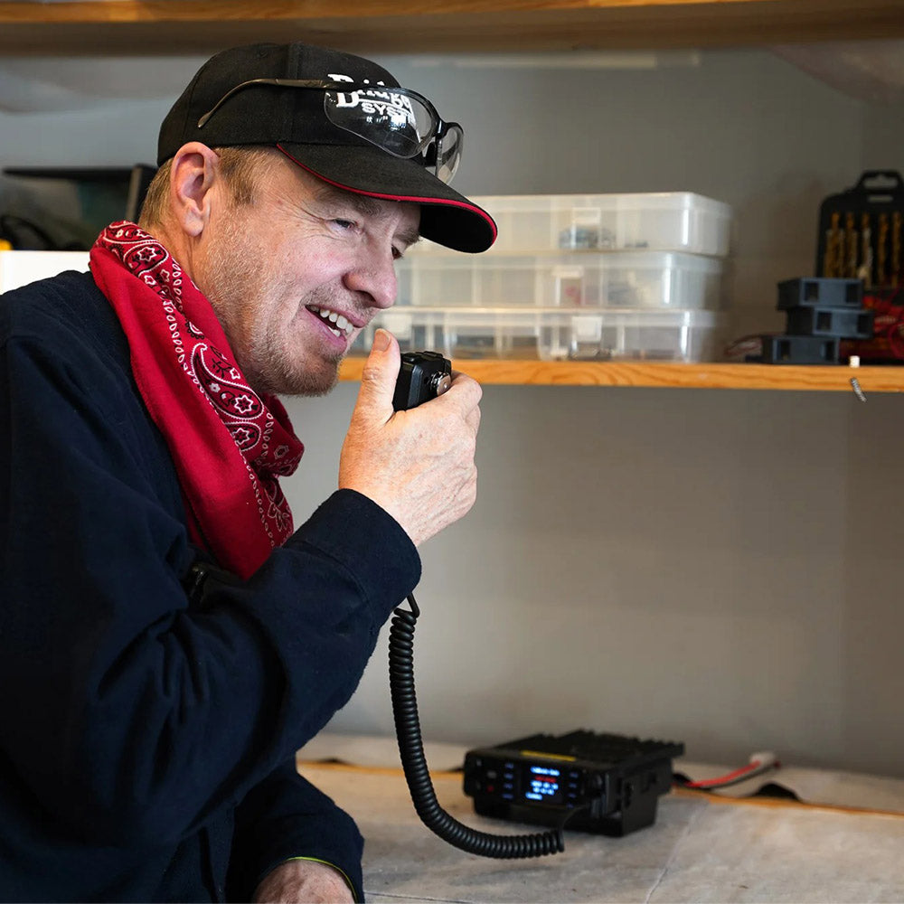 A man in a cap, red bandana, and glasses speaks into a handheld mic connected to an AnyTone 578UVIII PLUS Tri-Band Mobile Radio Freedom Bundle with $97 Training Course FREE! in a workshop with shelves and plastic boxes behind him.