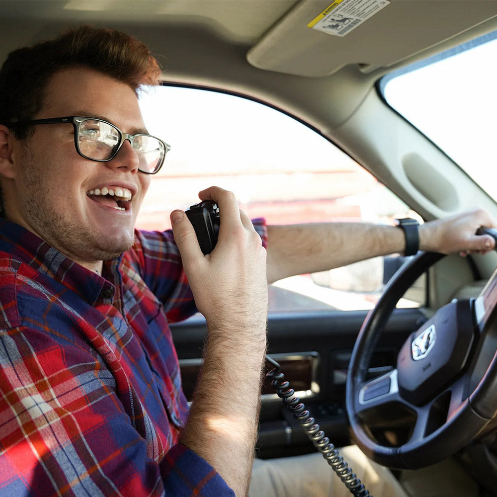 A smiling man wearing glasses and a red plaid shirt sits in the driver's seat of a car, speaking into the AnyTone 578UVIII PLUS Tri-Band Mobile Radio Freedom Bundle with FREE $97 Training Course.