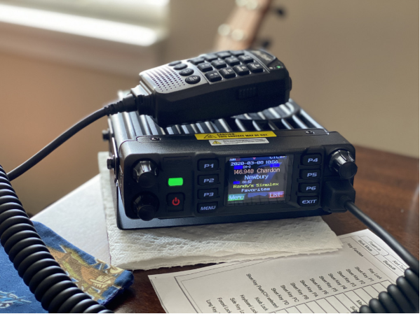 An AT-D578UVIII Plus Commercial Mobile Radio (GPS+BT) with various buttons and a small screen displaying frequencies and text sits ready for use. A handheld microphone rests on top of the radio, ideal for those who need DMR Repeater Roaming. A paper with handwritten notes is partially visible on the table, next to a cloth with a colorful design.