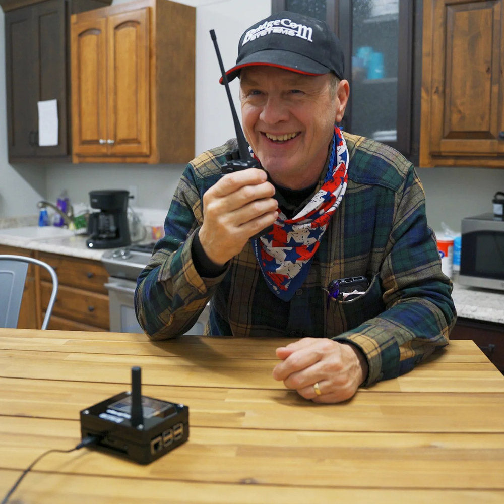 A smiling man in a plaid shirt and cap sits at a kitchen table, holding an AnyTone AT-D878UVII Plus radio. A SkyBridge MAX Hotspot rests nearby. Kitchen cabinets and appliances are visible in the background.