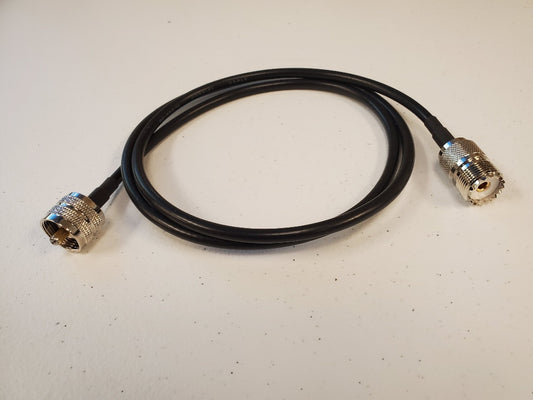 A 3' RG58 jumper cable with a PL259 male connector on one end and an SO239 female connector on the other is coiled in a loose circle and placed on a white surface, typically used for transmitting signals in various electronic devices.