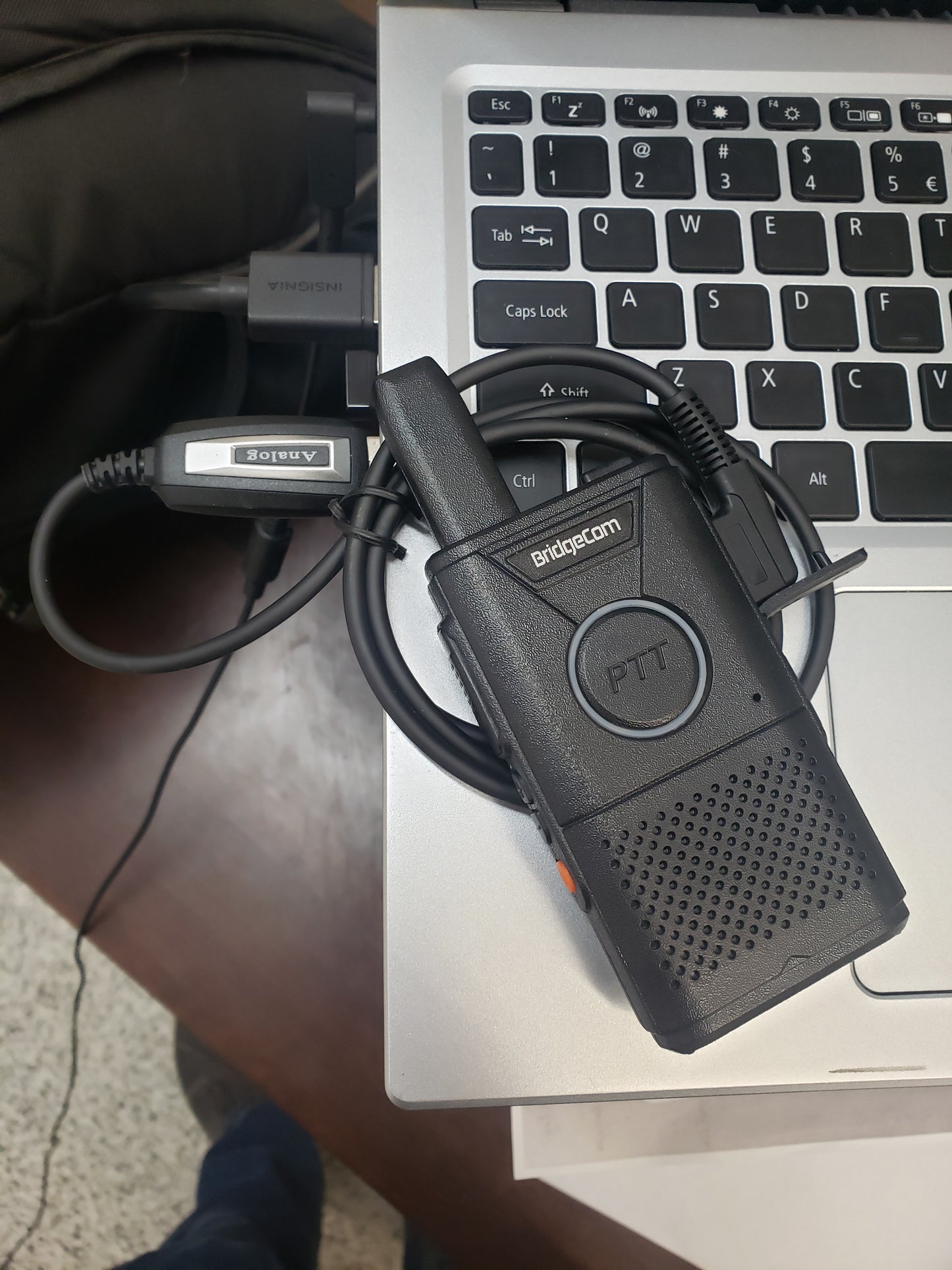 A BridgeCom Buddy Programming Cable with a coiled wire and earpiece is placed on the corner of an open laptop keyboard; part of a backpack and a laptop charger appear in the background.
