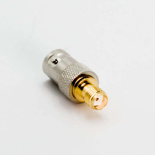 Close-up image of an SMA F to BNC F coaxial adapter, featuring a BNC female connector on one end and an SMA male connector on the other. The adapter, designed for an AnyTone handheld, boasts a silver metallic finish with a knurled middle section for grip and a gold-plated SMA end. It is placed on a plain white background.