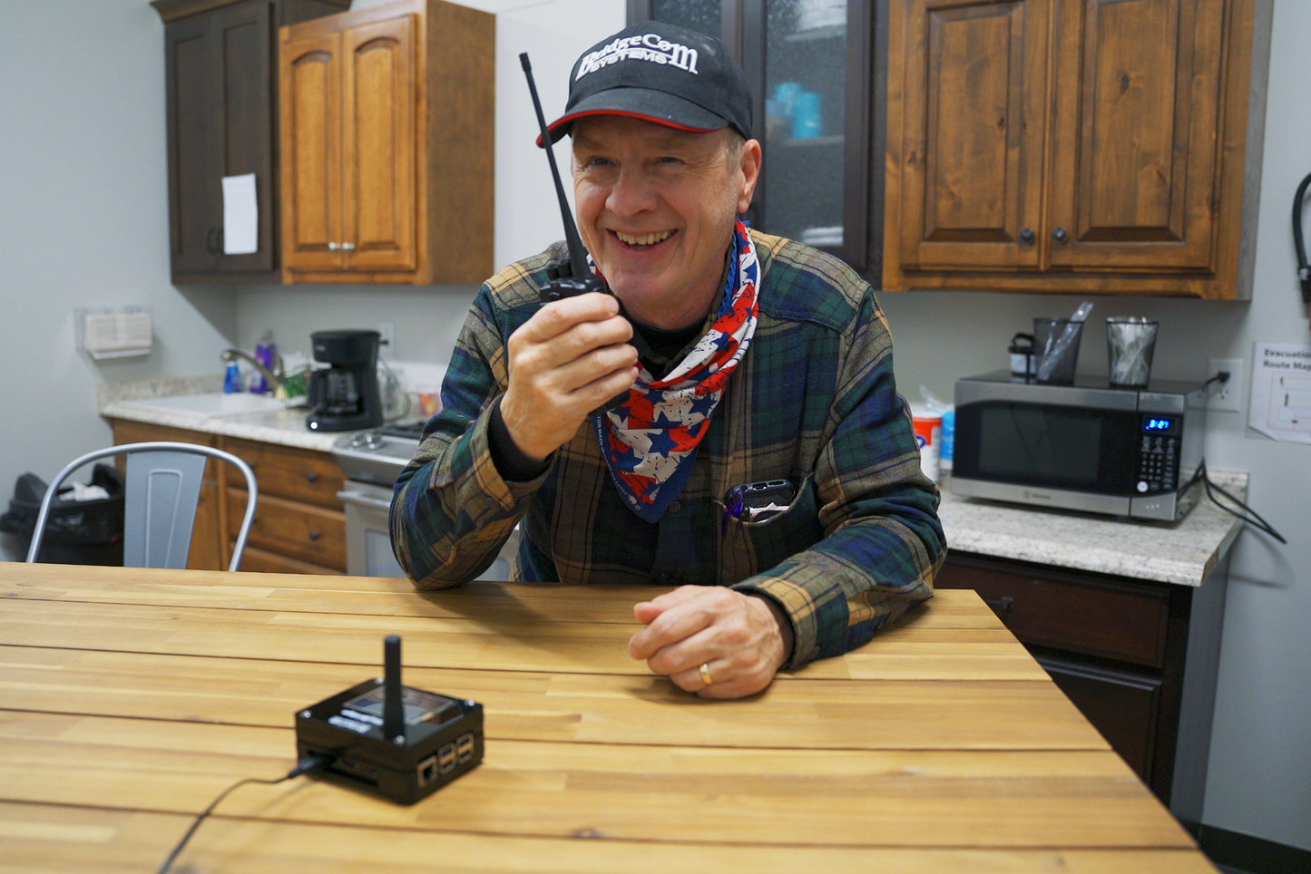 A man wearing a plaid shirt, cap, and bandana sits at a wooden table holding an Emergency Plug and Play Package with a $194 Training Course FREE! along with an antenna. He is smiling and appears to be speaking into the device. The background includes a kitchen area with wooden cabinets, a microwave, and a coffee maker.