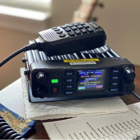 A black B-Stock AnyTone AT-D578UVIII Plus ham radio transceiver on the table features various buttons and a digital display showing frequencies, with a microphone resting on top, connected by a cord. B-stock papers and a cloth piece are placed under the radio, which is capable of receiving Air Band signals.