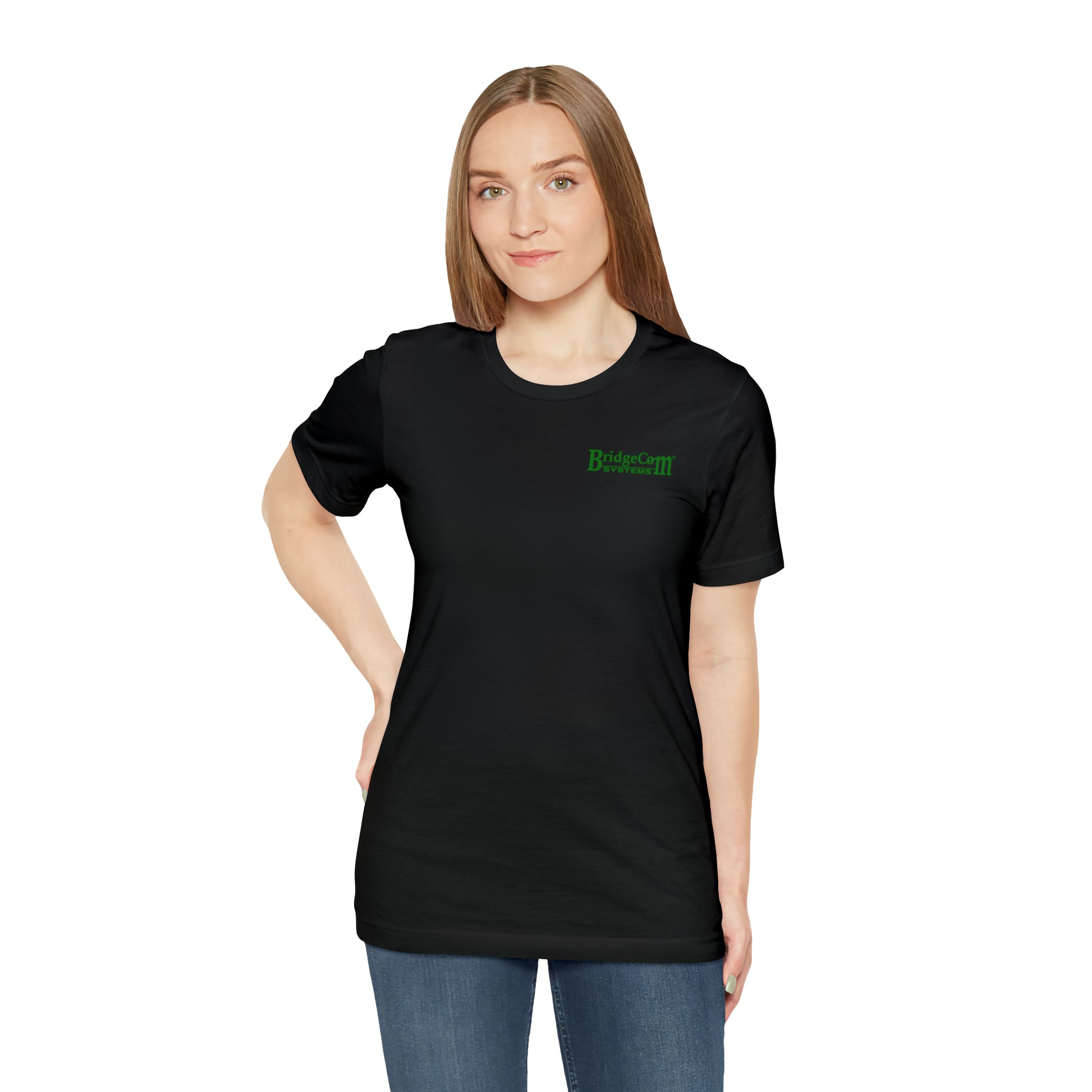 A person with long hair stands against a white background, wearing a Short Sleeve Tee - Small Logo in black with green text reading "BridgeCom Systems" on the upper left side. Made from 100% Airlume cotton, their left hand rests on their hip while their other arm is relaxed by their side. They are also wearing blue jeans.