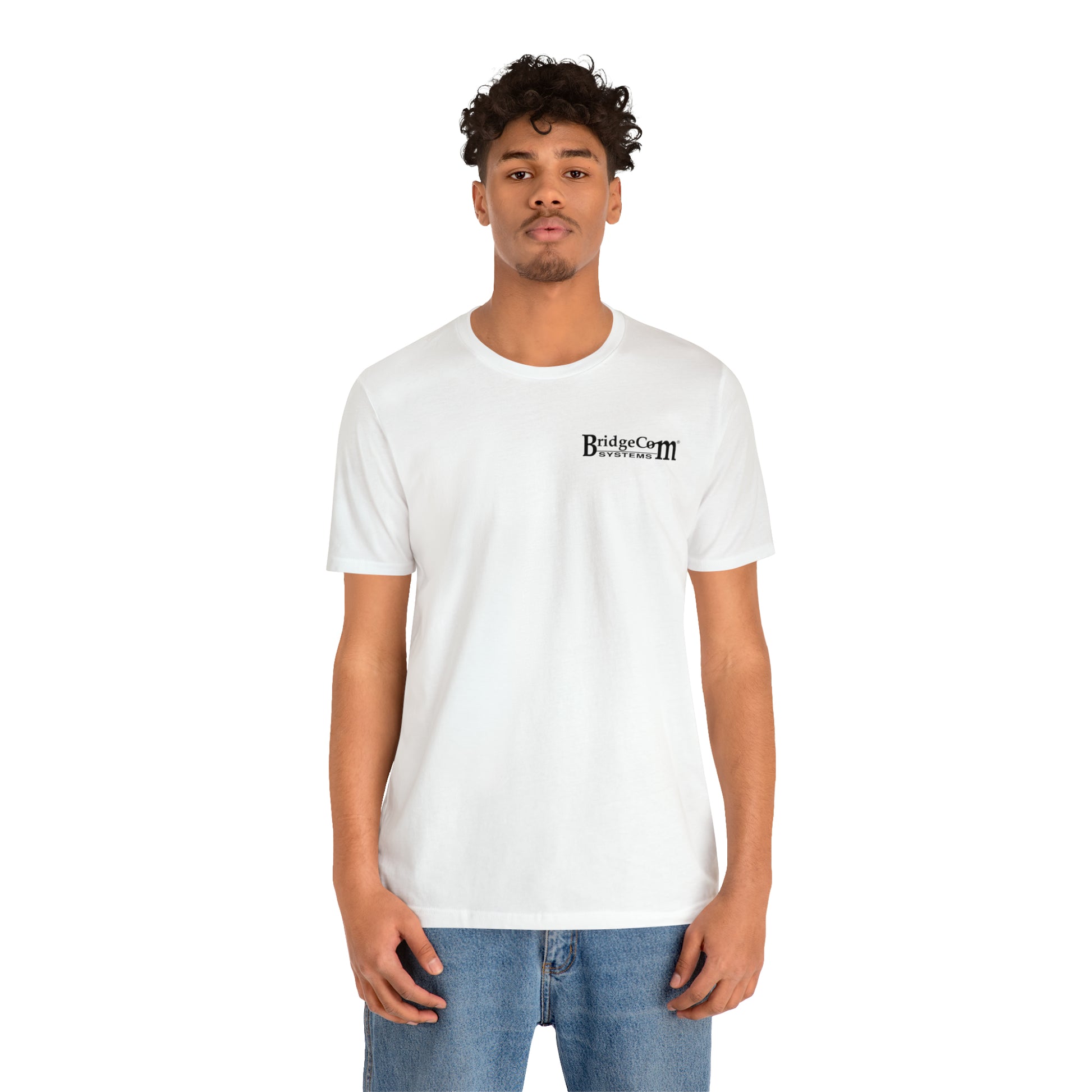 A person with short curly hair stands against a white background, wearing a BridgeCom Systems Short Sleeve Tee - Small Logo made of soft Airlume combed cotton, paired with blue jeans.