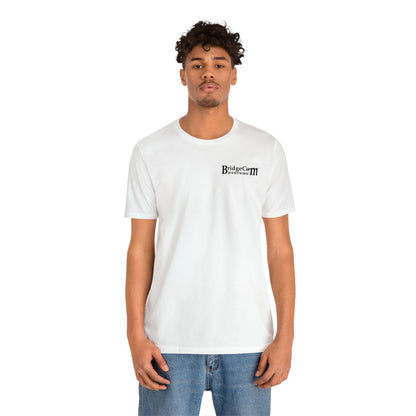 A person with short curly hair stands against a white background, wearing a BridgeCom Systems Short Sleeve Tee - Small Logo made of soft Airlume combed cotton, paired with blue jeans.