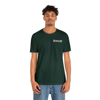 A person with curly hair is wearing a dark green Short Sleeve Tee - Small Logo and blue jeans. They are standing against a plain white background, facing the camera directly with a neutral expression.