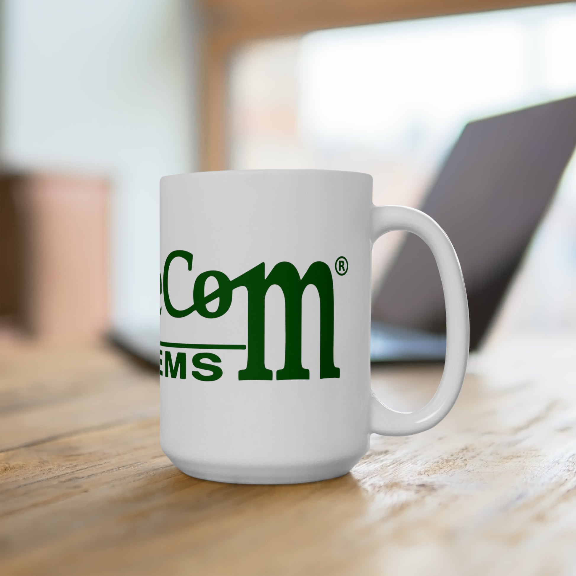 A customizable BridgeCom Ceramic Mug 15oz with green text sits on a wooden table. In the background, a slightly blurred laptop is partially visible. The 15-ounce mug features a partially obscured brand logo with the letters "MS" prominently displayed.