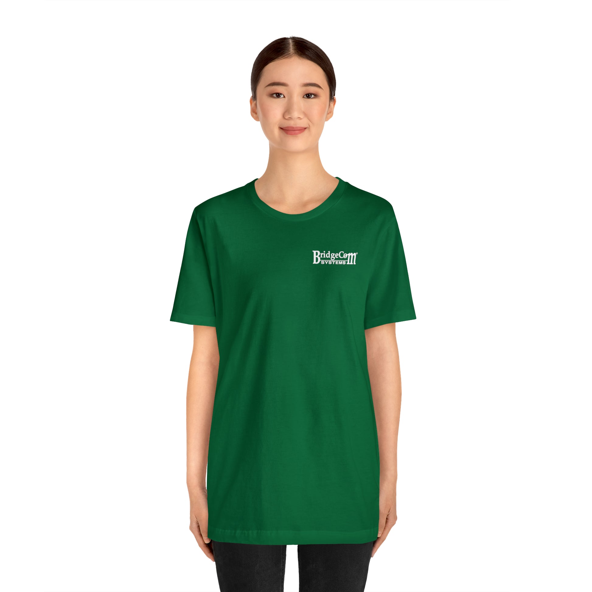 A young woman stands smiling, wearing a green Short Sleeve Tee - Small Logo made of soft Airlume combed cotton. The T-shirt features a small white logo on the top left that reads "Bird & Cat Doormat." She has dark hair tied back, and her hands are by her sides. The background is plain and white.