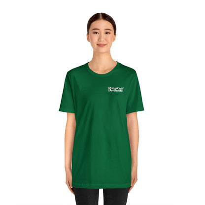 A young woman stands smiling, wearing a green Short Sleeve Tee - Small Logo made of soft Airlume combed cotton. The T-shirt features a small white logo on the top left that reads "Bird & Cat Doormat." She has dark hair tied back, and her hands are by her sides. The background is plain and white.
