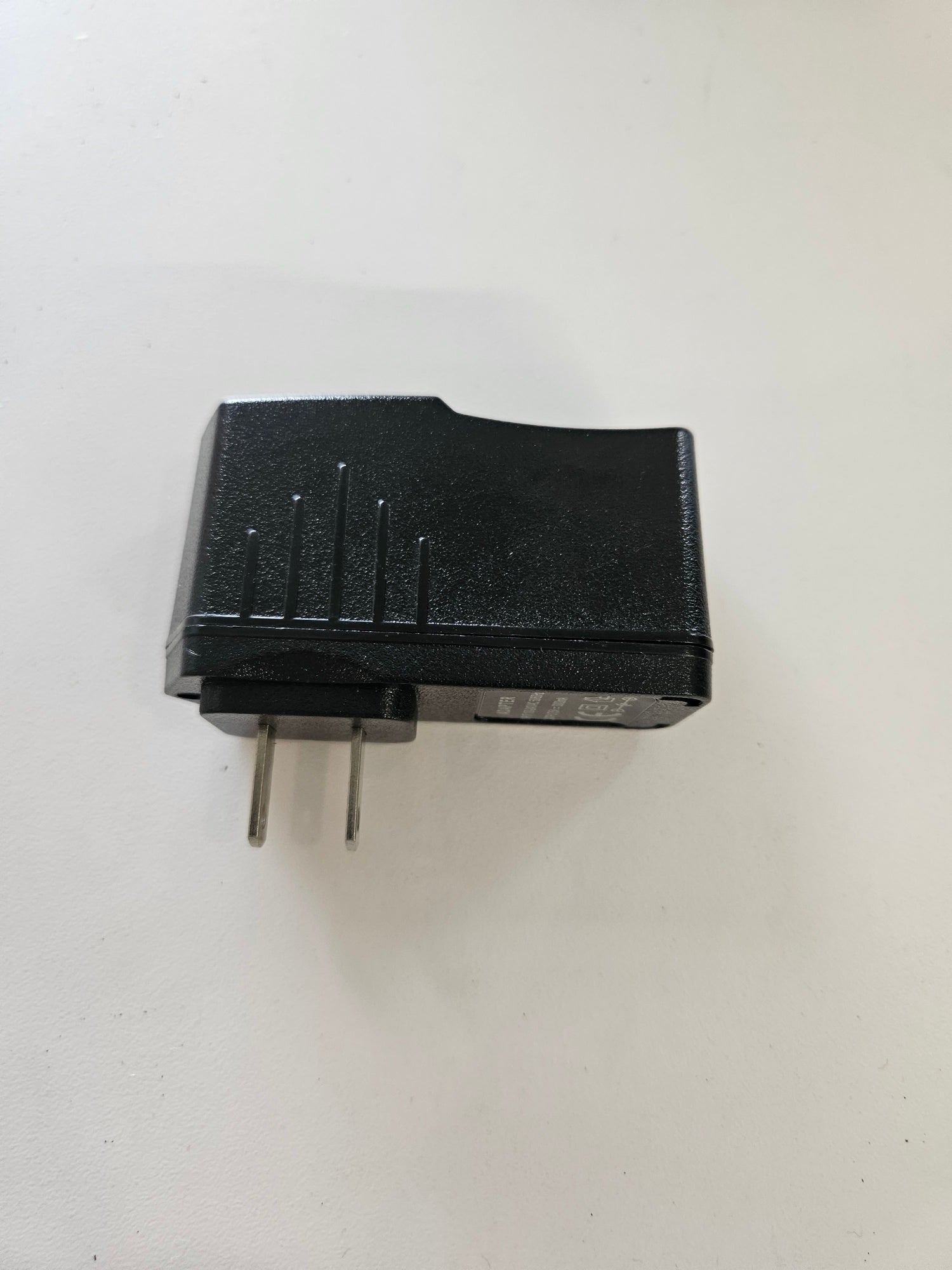 A black 5V 3A Skybridge Power Adapter-Wall Charger with two metal prongs is lying on a white surface. The charger features a slightly textured finish with a few ridges on one side.