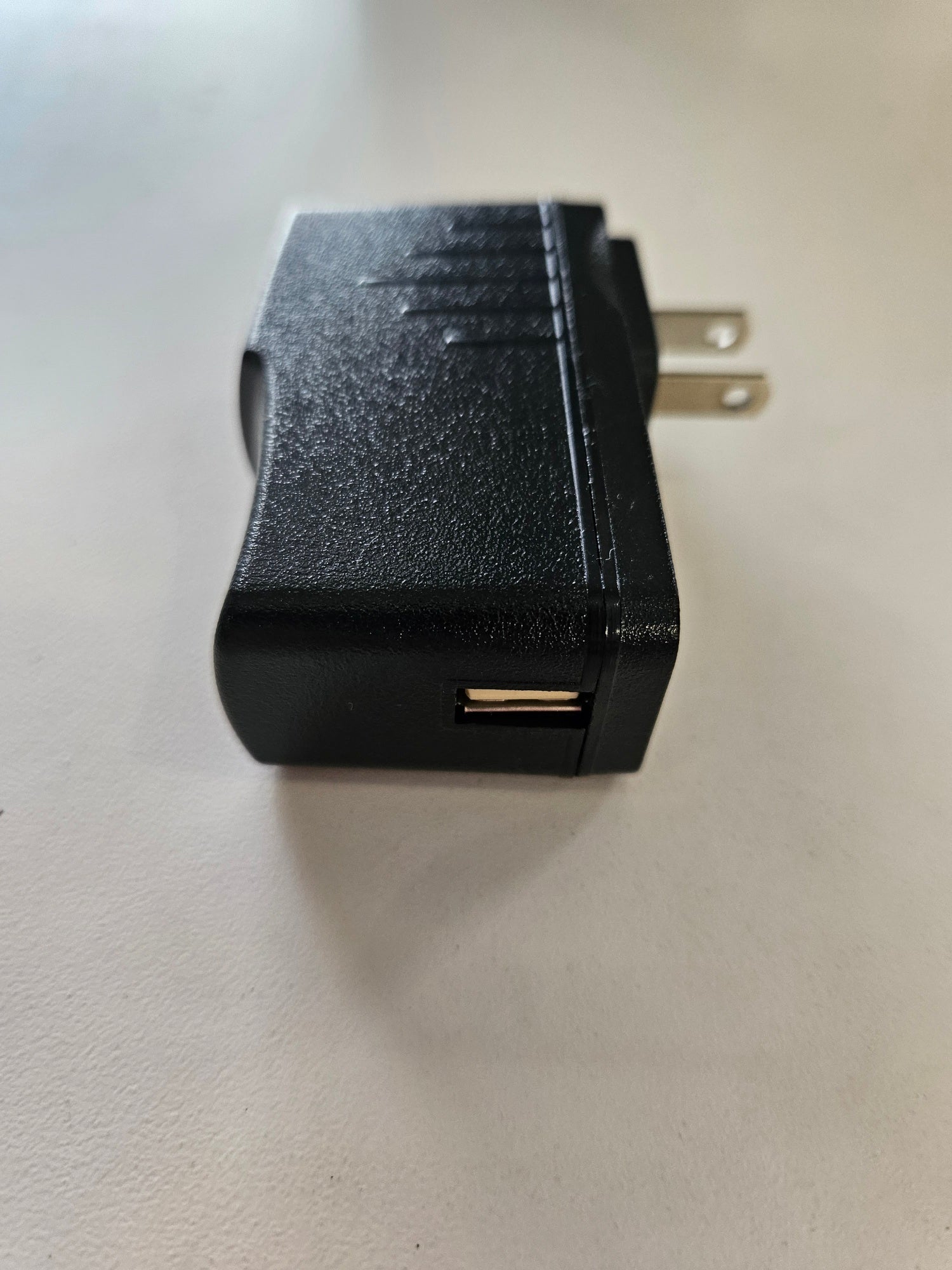 A black 5V 3A Skybridge Power Adapter - Wall charger with a textured surface and two metal prongs for plugging into an electrical outlet, lying on a light-colored surface. The USB port is visible on the side of the charger.