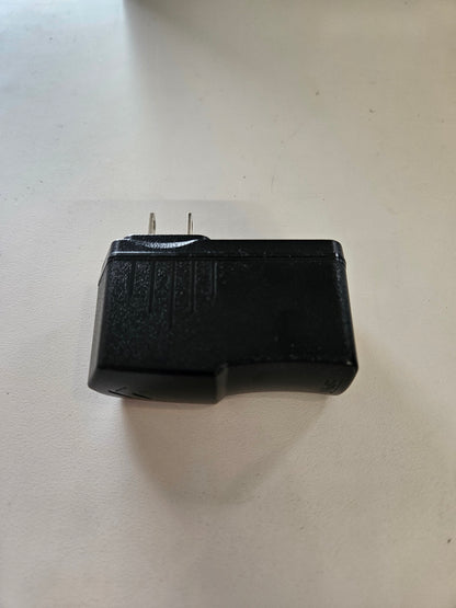 A black, rectangular 5V 3A Skybridge Power Adapter – Wall charger with upward-facing prongs lies on a flat, white surface. Its casing is adorned with textured lines and grooves, which give it a slightly worn or used appearance.