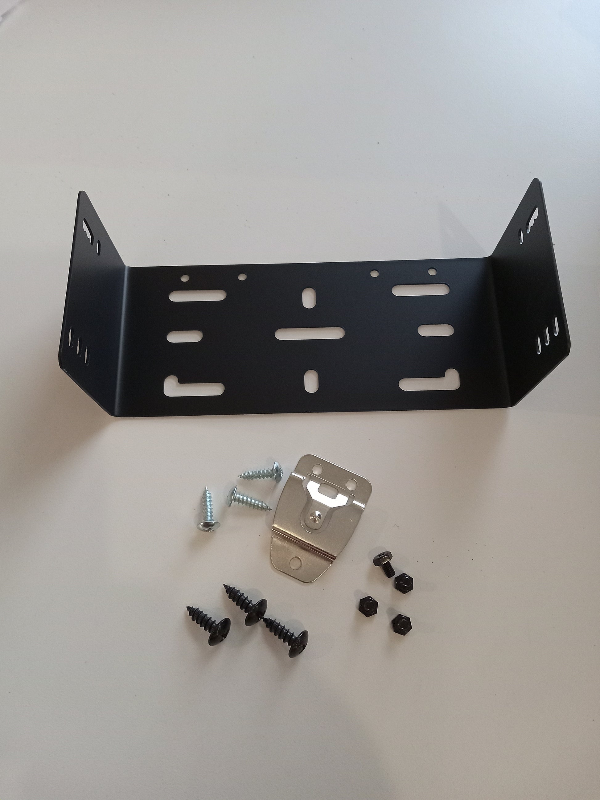 On a white surface lies the BCM Mounting Bracket with Hardware, a black metal bracket featuring various slots and holes. Accompanying it are several screws, nuts, and a small metal piece with a pre-drilled hole in the center. This setup incorporates the versatile BCM-220 model for enhanced compatibility.