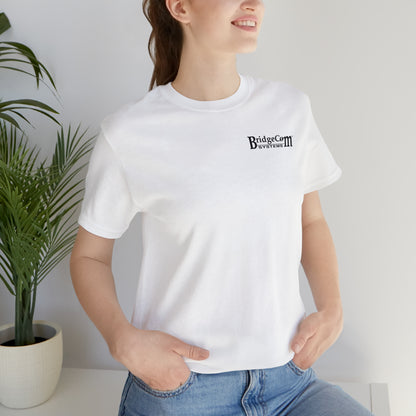 A person wearing a Short Sleeve Tee - Small Logo with the BridgeCom Systems logo and "BridgeCon October 2023" printed on the chest is standing next to a potted plant with green leaves, smiling slightly. The soft Airlume combed and ringspun cotton shirt makes them look effortlessly comfortable, one hand in their blue jeans pocket.