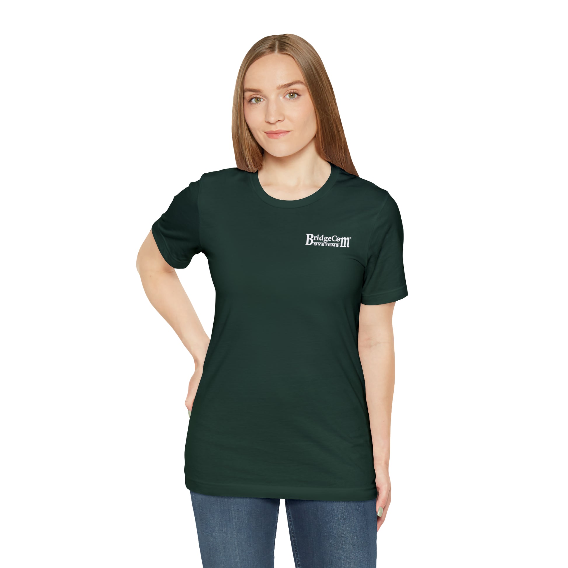 A woman with long, straight hair is wearing a dark green "Short Sleeve Tee - Small Logo" shirt with the "BridgeCom Systems" logo on the left side of her chest. She stands with one hand on her hip and has a neutral expression. She is also wearing blue jeans, set against a plain white background, emphasizing her connection to communication.