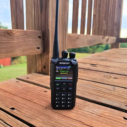 A black BridgeCom Maverick Dual Band DMR/Analog Ham Radio with an antenna stands upright on a wooden deck, with green grass and a partly cloudy sky in the background.