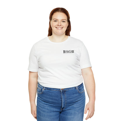 A smiling person with long red hair stands against a white background. They are wearing a Short Sleeve Tee - Small Logo proudly displaying "BridgeCom Systems," along with blue jeans, embodying the spirit of efficient communication.