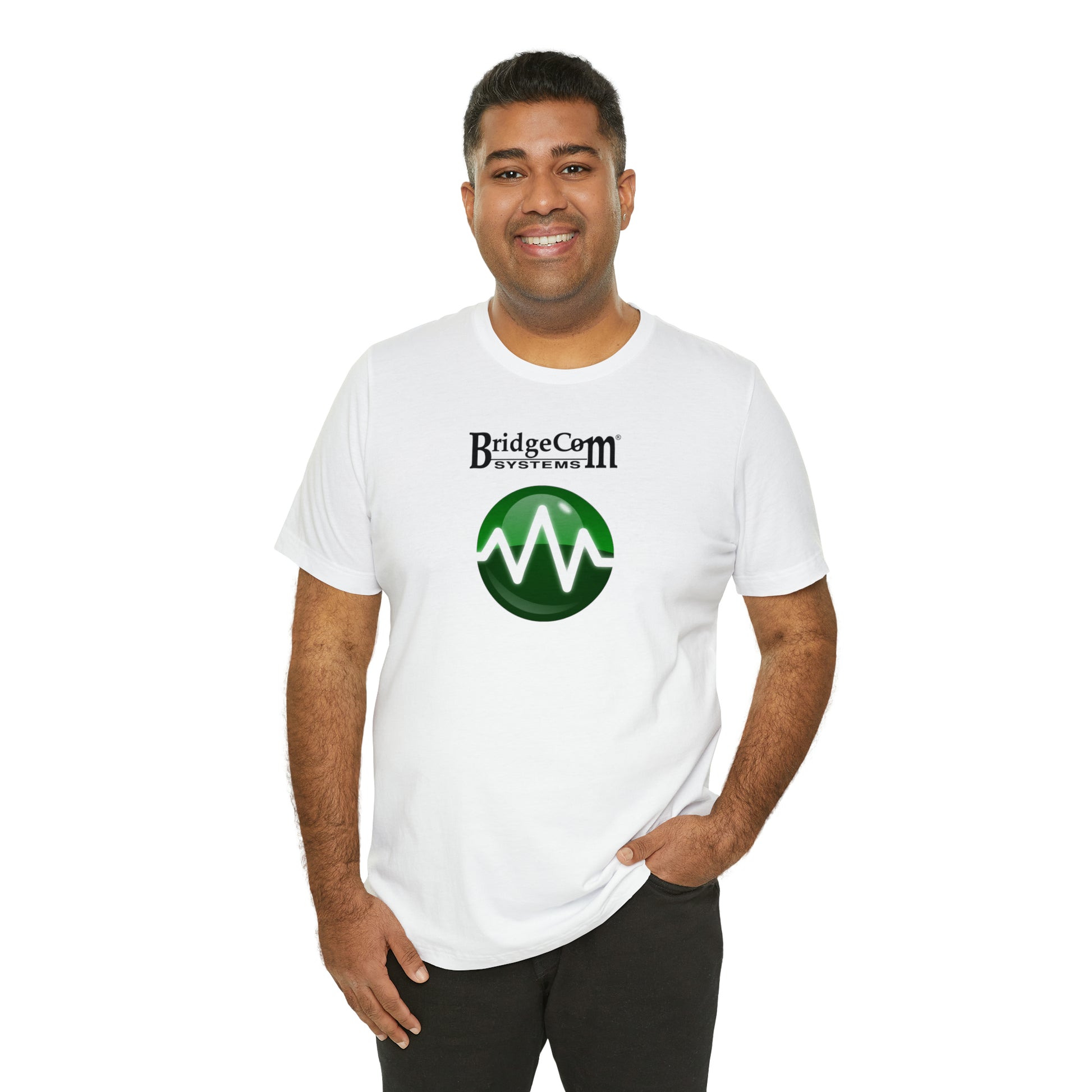 A smiling person stands with one hand in their pocket, proudly wearing a white "Unisex Short Sleeve BridgeCom Tee - Alternate Logo" made of soft Airlume cotton. The T-shirt features the "BridgeCom Systems" logo with a green circle and wave pattern, all set against a plain white background.