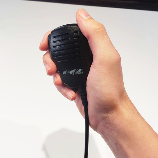A hand holding a black BridgeCom BCS-200 Water Resistant Speaker Mic with a coiled cable extending from the bottom. The microphone, compatible with a Kenwood Style plug, features wind porting for clear audio, a push-to-talk button on the left side, and a small speaker grille on the front. The background is plain and light-colored.