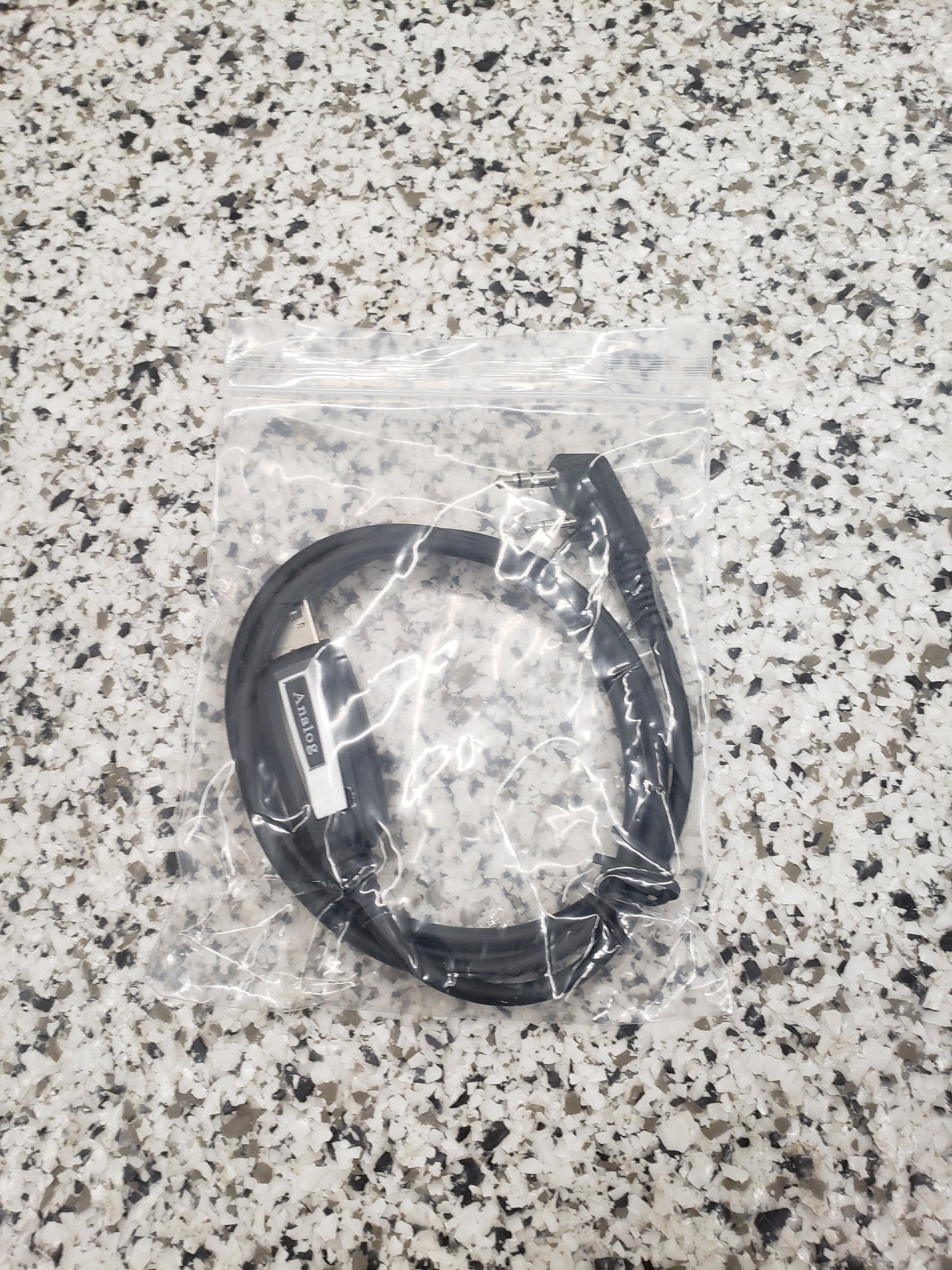 The BridgeCom Buddy Programming Cable, in black and packaged in a clear ziplock bag on a gray granite countertop, is perfect for programming your Buddy FRS handheld radio.
