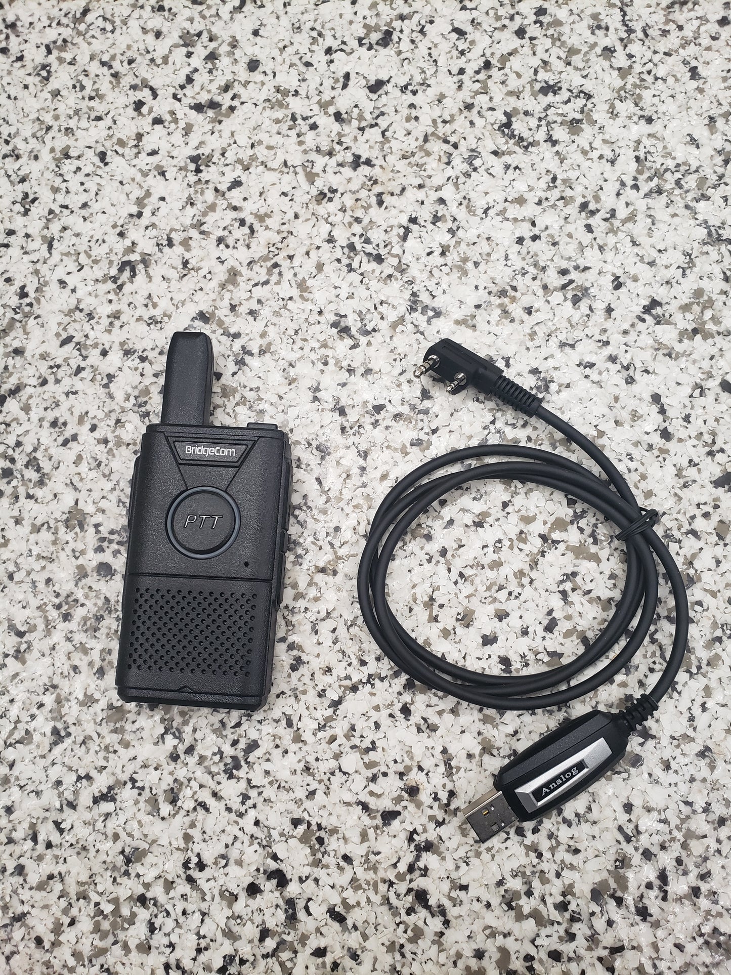 A black Buddy FRS handheld radio and the BridgeCom Buddy Programming Cable rest on a speckled gray countertop. The radio displays a "PTT" button, while the cable features a USB connector and right-angle plug for easy programming.